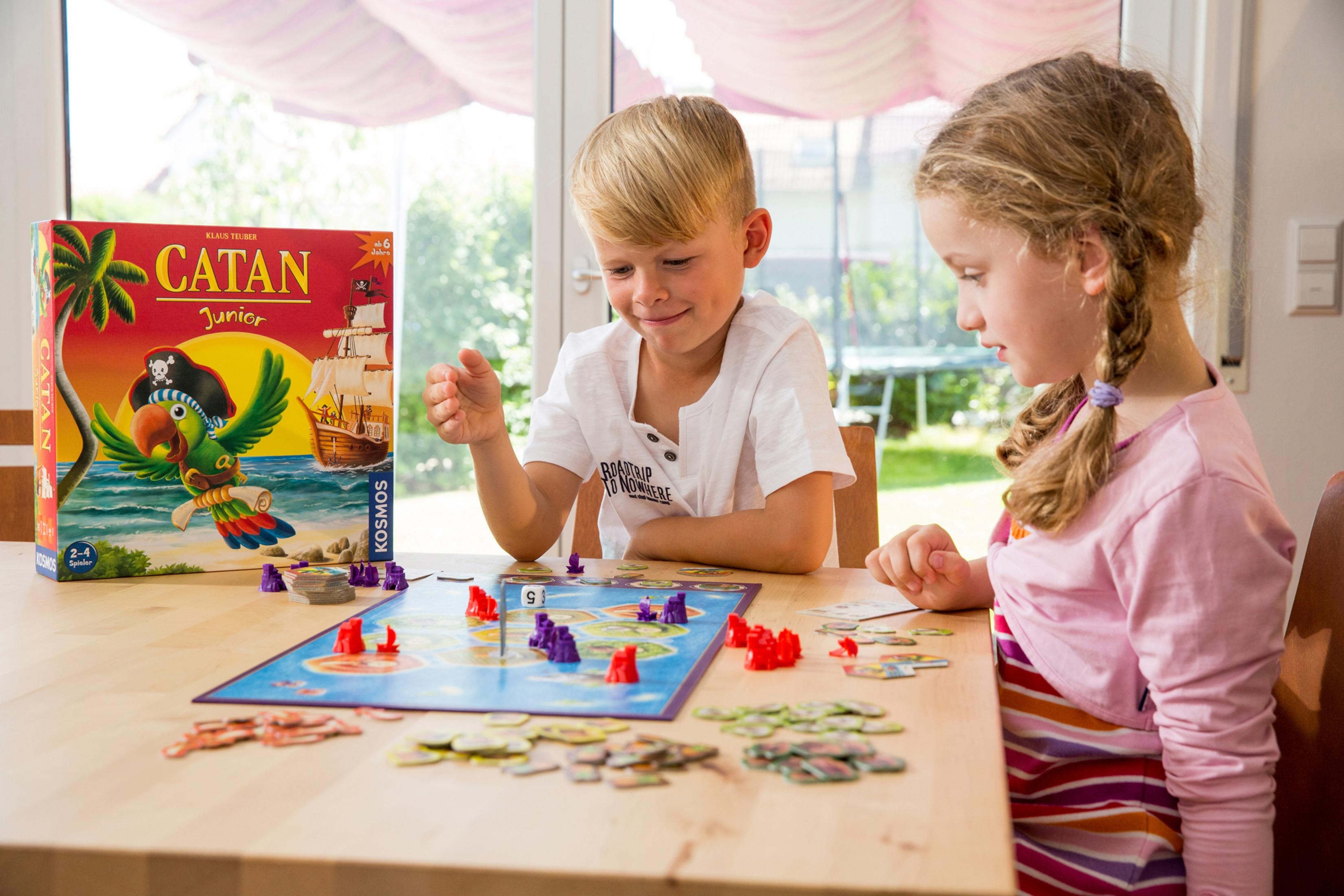 Zwei Kinder spielen Catan Junior Brettspiel an einem Holztisch, lächeln und interagieren.