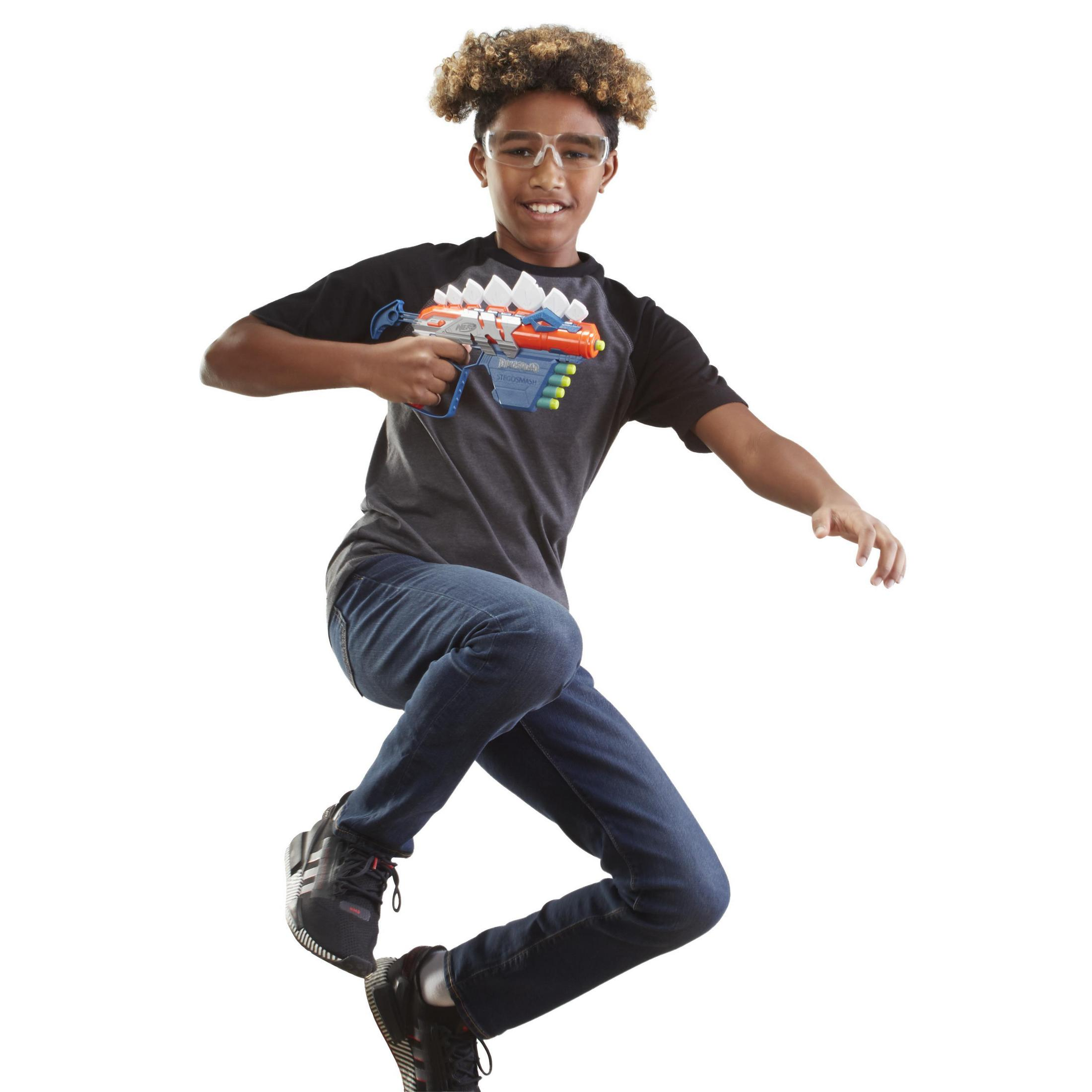 Junge springt, hält Spielzeugpistole, trägt Brille und Jeans, weißer Hintergrund.