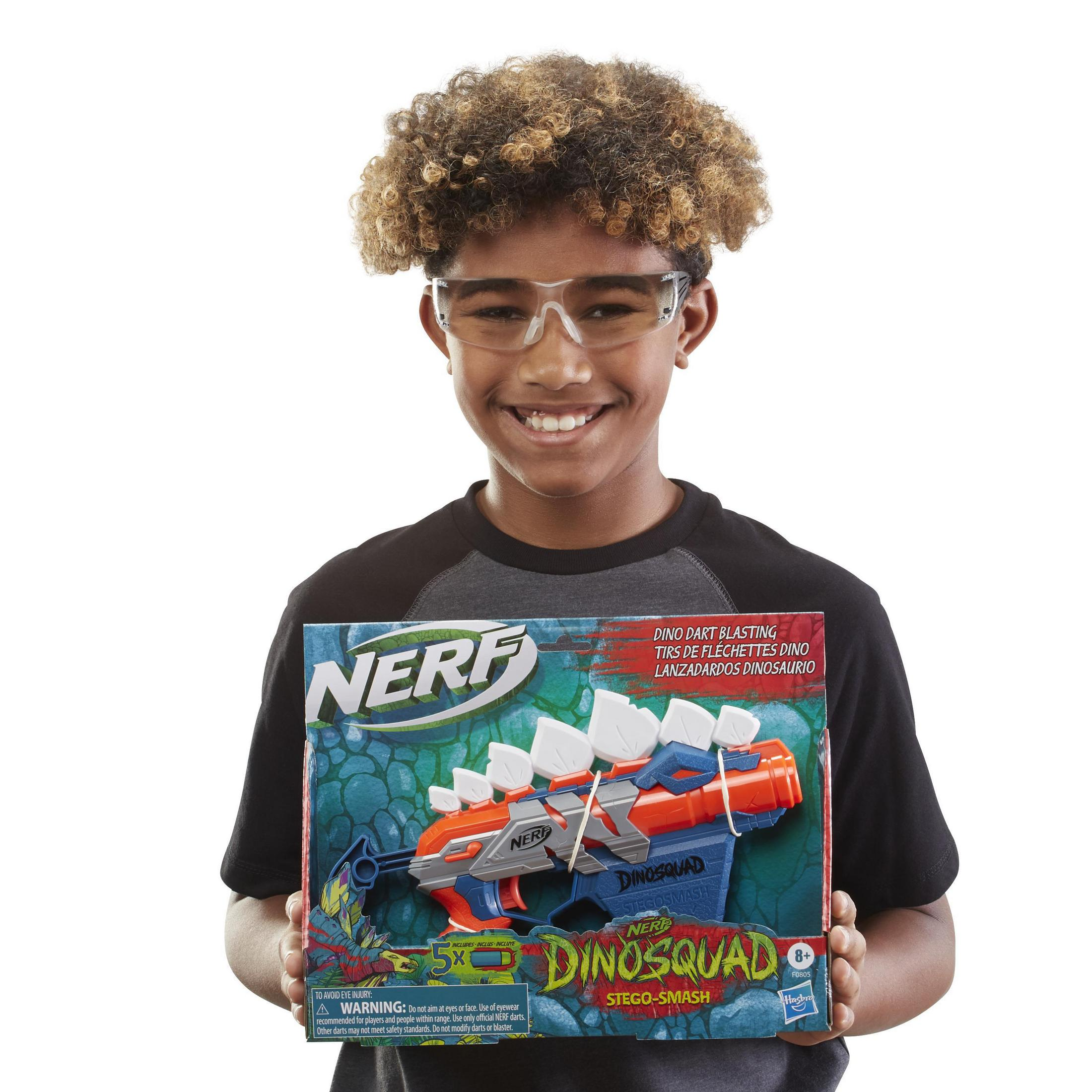 Junger Junge hält ein Nerf DinoSquad Spielzeug, lächelt, trägt eine Brille, auf weißem Hintergrund.
