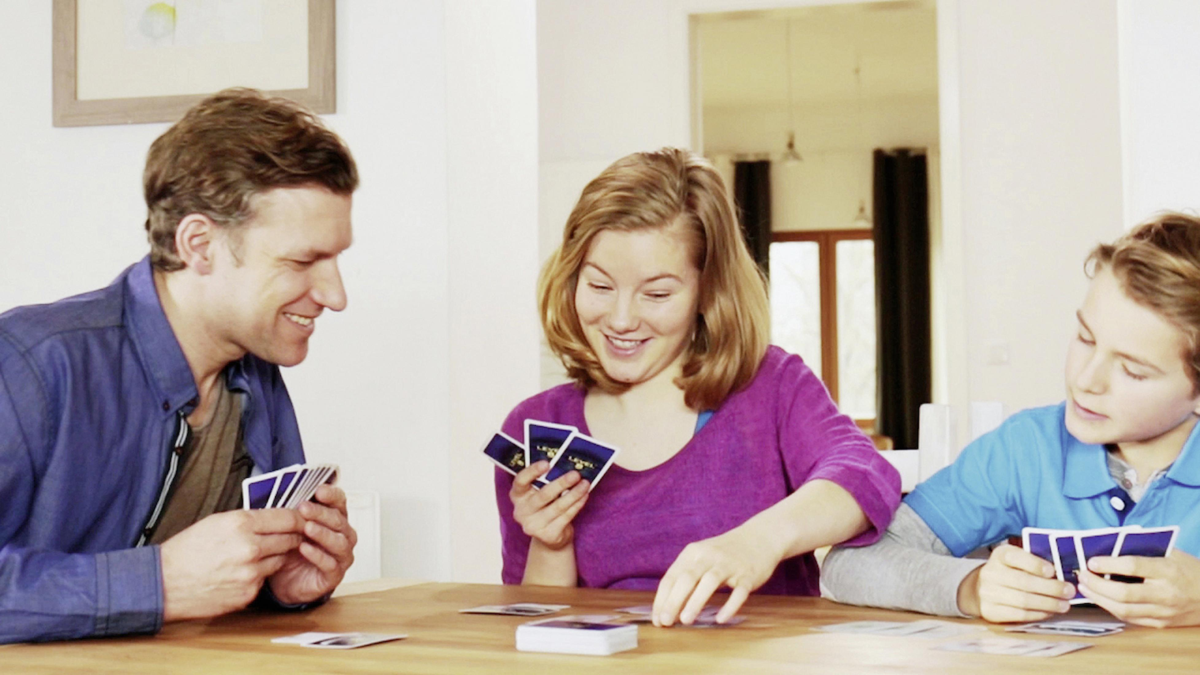 Rodzina grająca w karty przy stole, uśmiechnięta. Mężczyzna, kobieta i chłopiec są na zdjęciu.