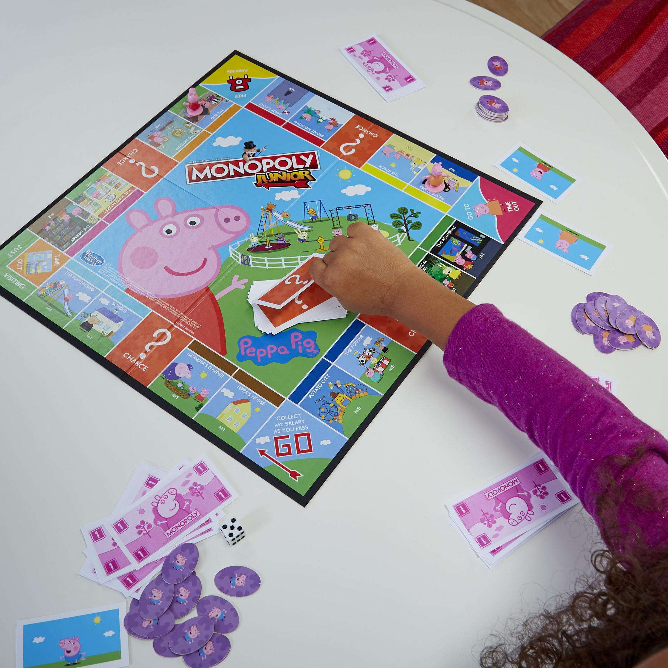 Gra planszowa Monopoly Junior z motywem Peppy Pig. Ręka ma za chwilę wylosować kartę. Widoczna plansza i karty.