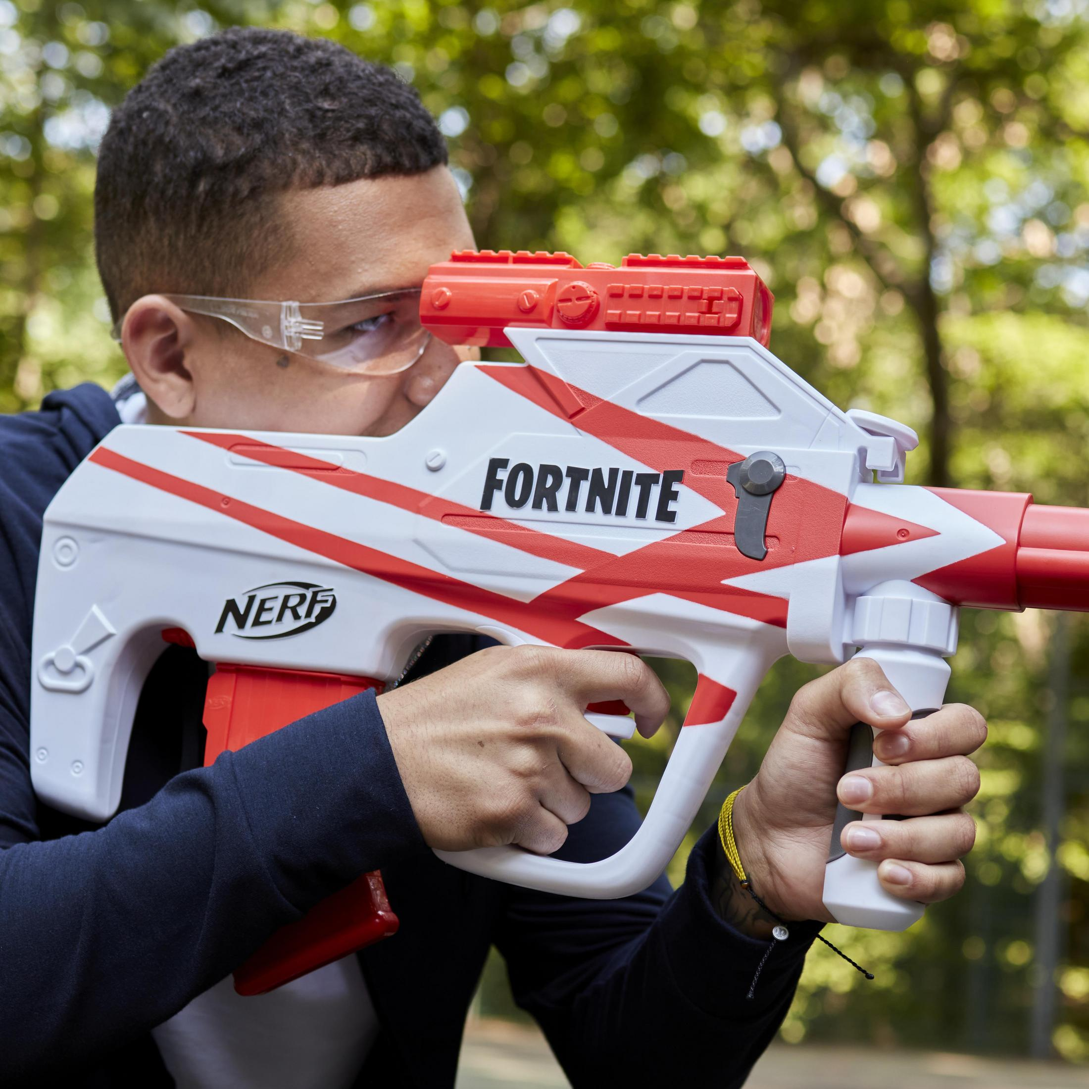 Mężczyzna w okularach celuje biało-czerwoną bronią Nerf Fortnite. Tło to rozmyty las.