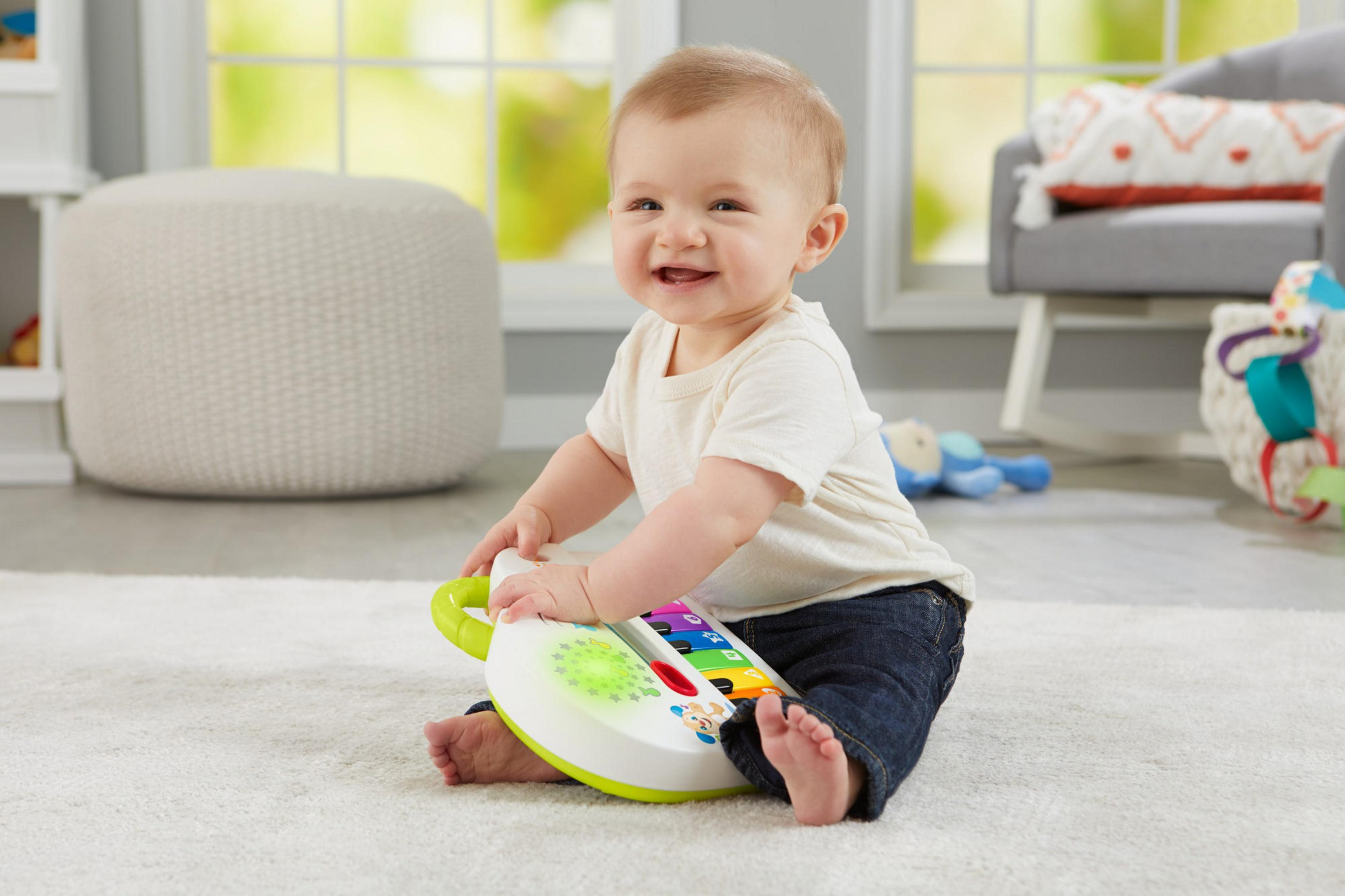 Ein Baby sitzt auf einem Teppich und spielt mit einem Spielzeugklavier. Das Baby lächelt.
