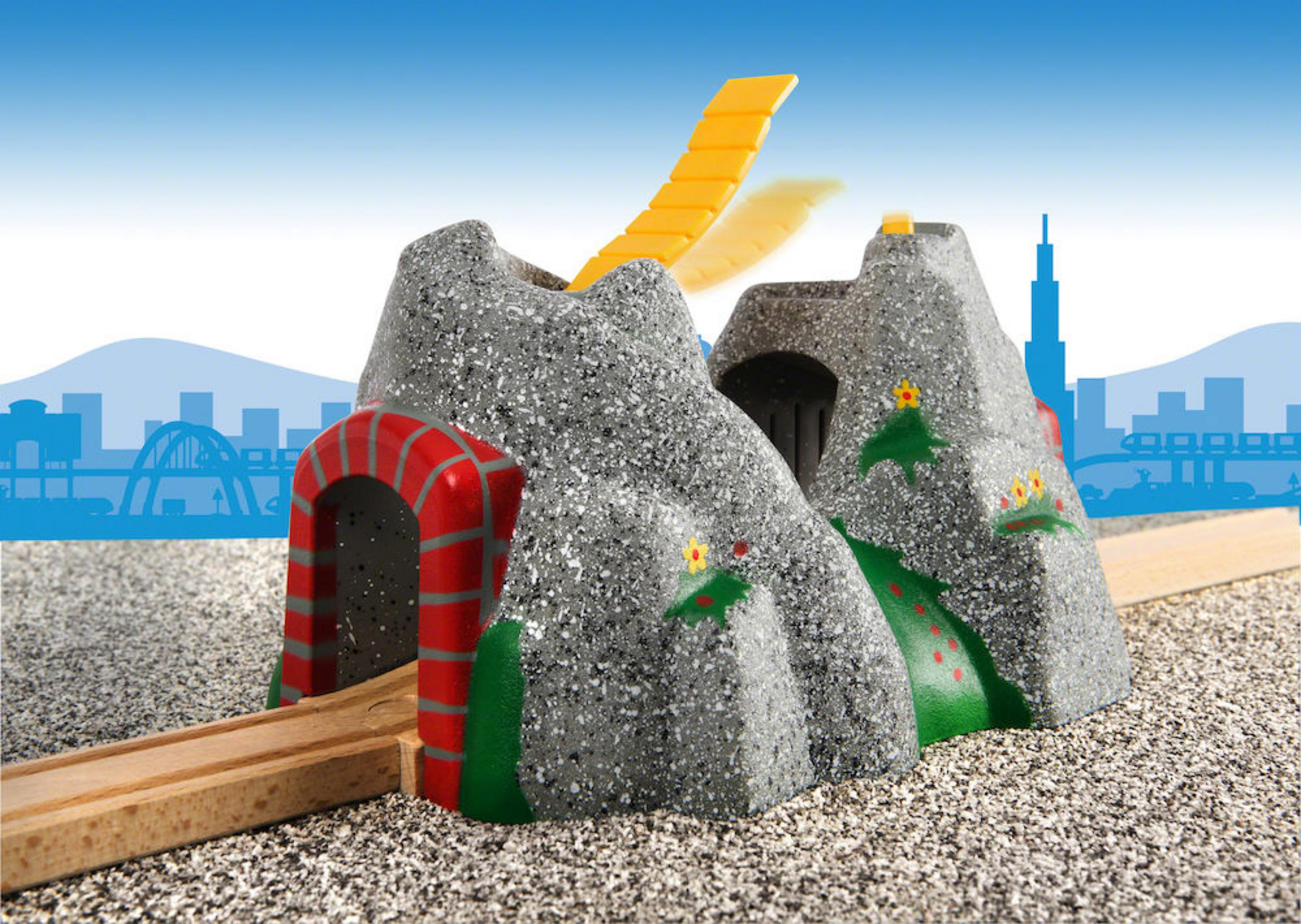 Eine Spielzeug-Zugstrecke mit grauem Bergtunnel, gelber Rampe und Stadtsilhouette.