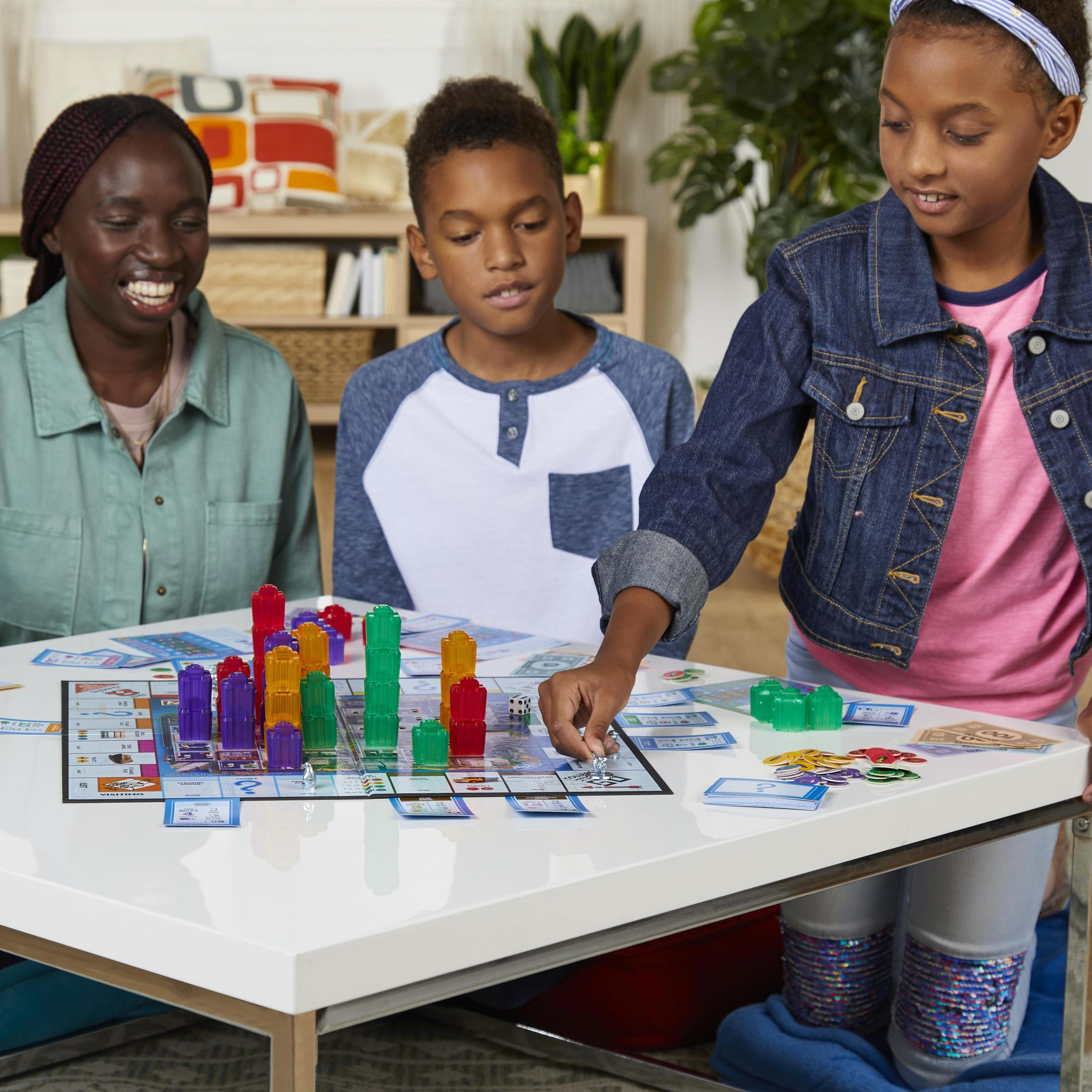 Drei Kinder spielen Monopoly mit Spielbrett, Häusern, Würfeln und Geld auf einem weißen Tisch.