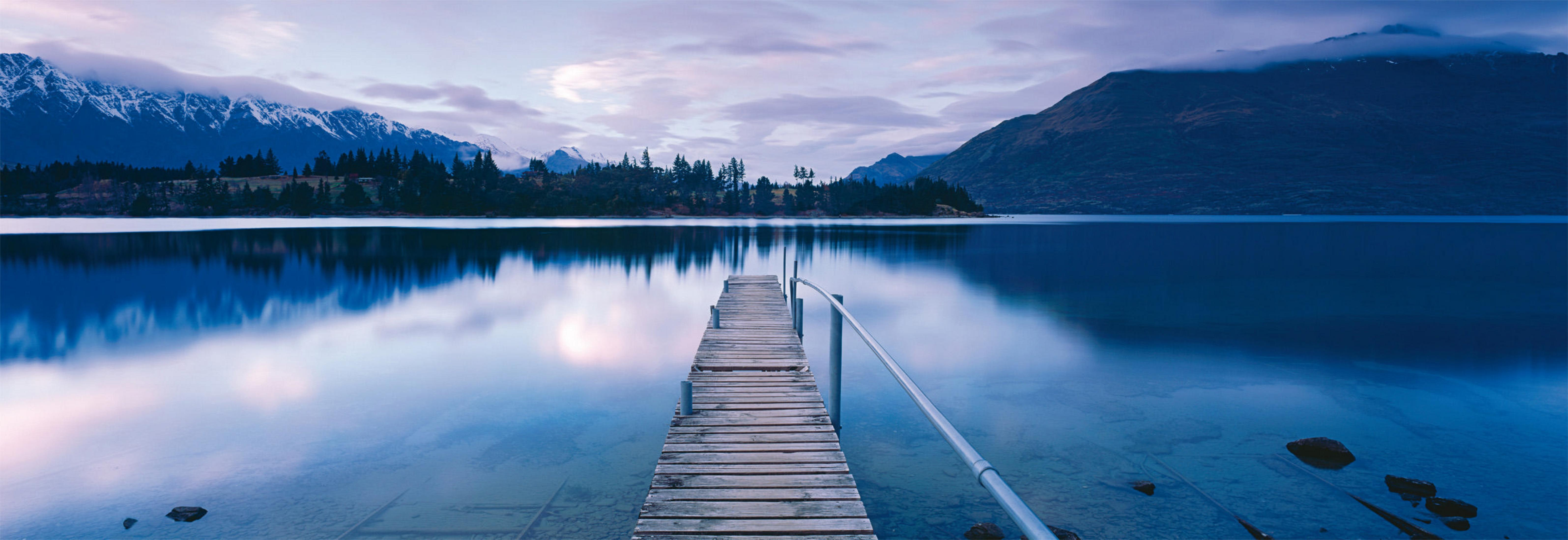 Vista panoramica del lago, un molo di legno si estende nell'acqua calma, montagne e cielo nuvoloso sullo sfondo.