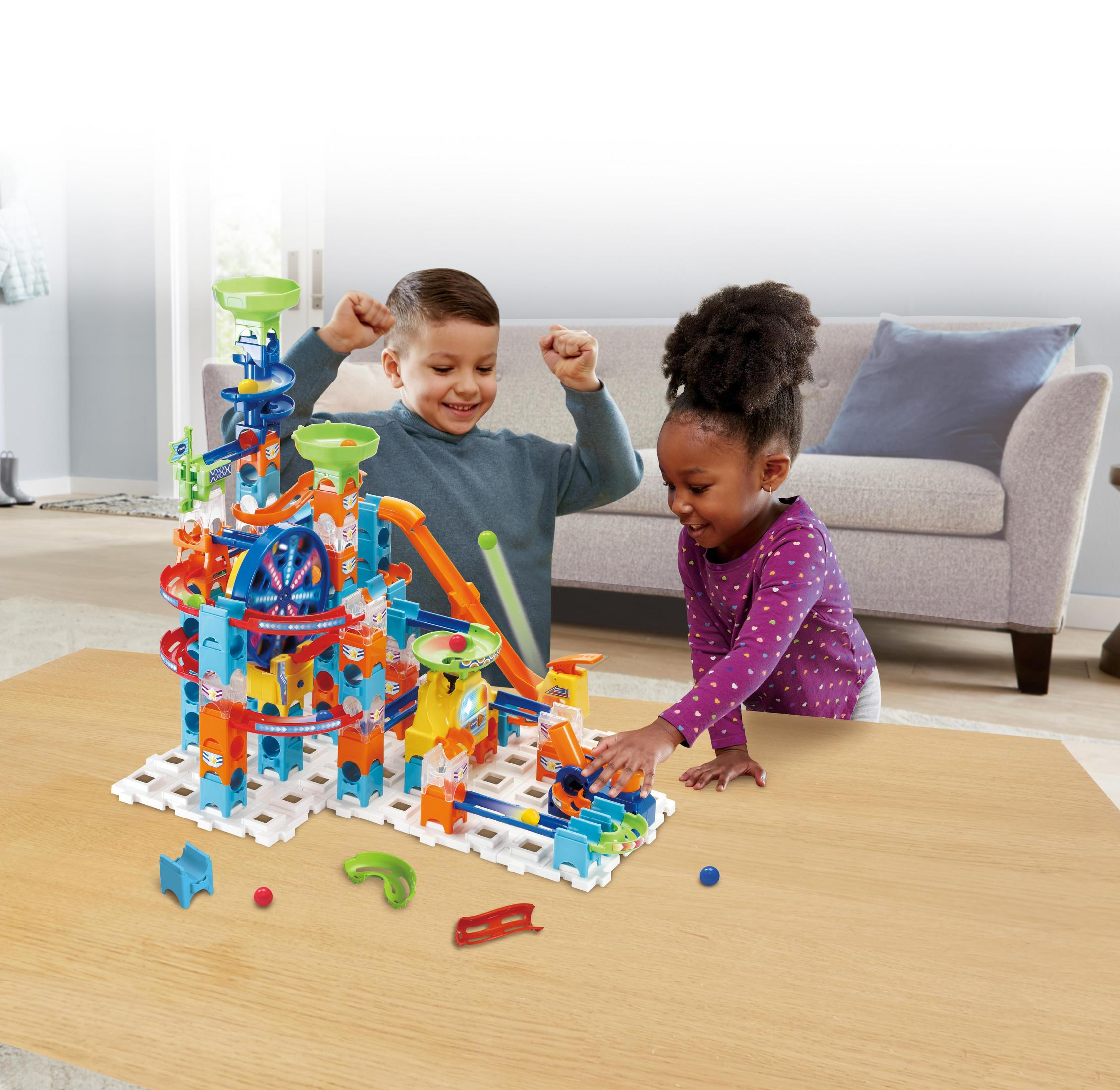 Zwei Kinder spielen mit einem Murmelbahnspielzeug auf einem Tisch. Ein Sofa ist im Hintergrund sichtbar.
