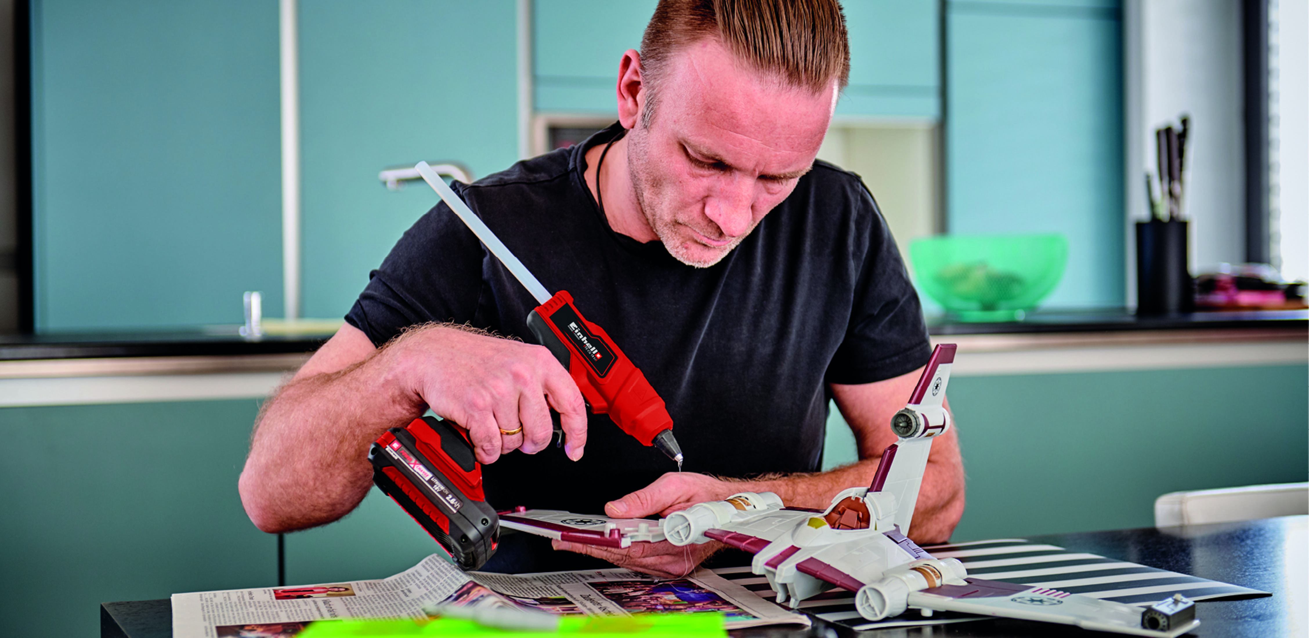 Mężczyzna używający pistoletu do klejenia na modelu Star Wars na stole z gazetą.