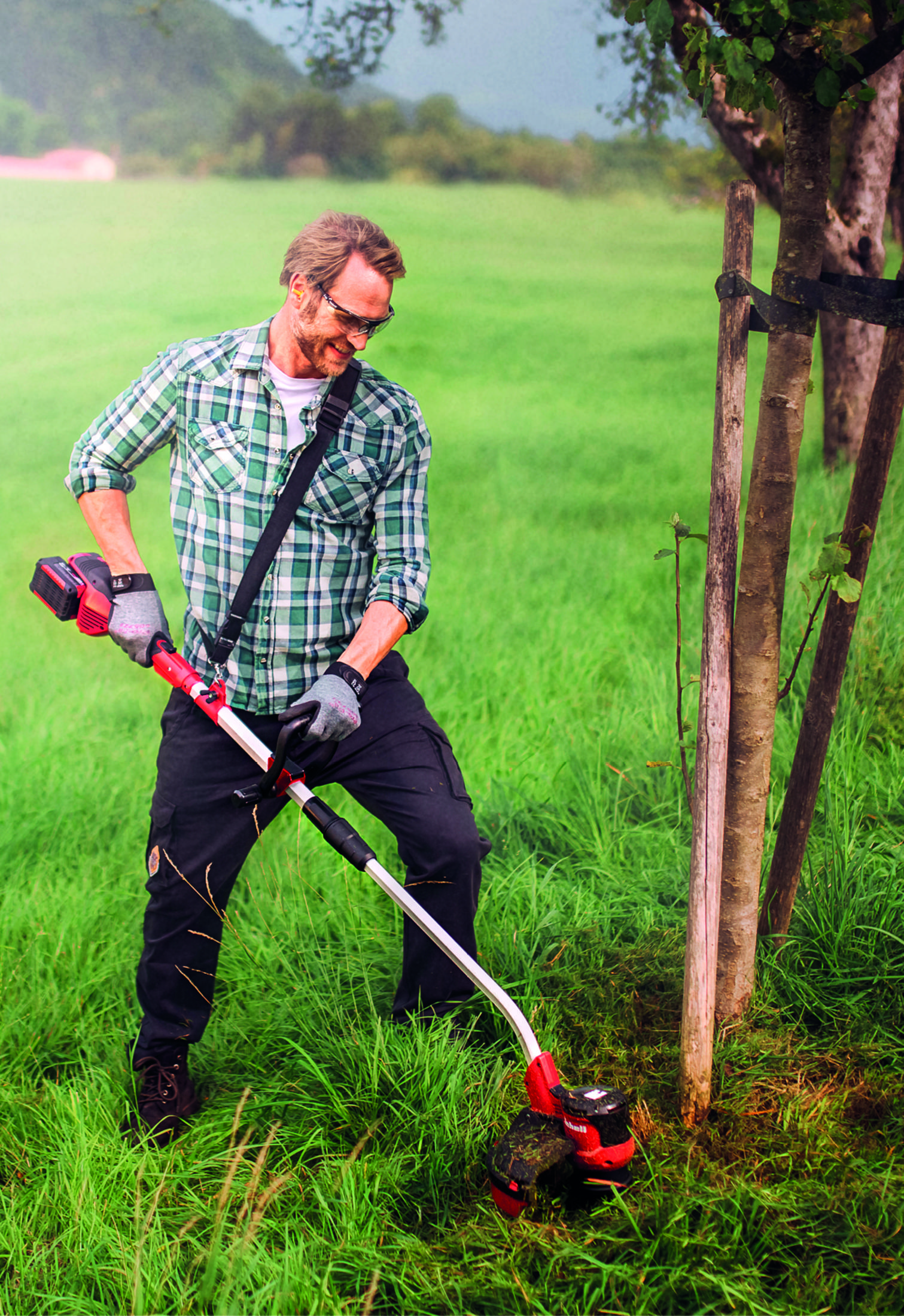 Hombre usando un cortasetos rojo y negro en césped, cerca de árboles. Usa gafas y equipo de protección.