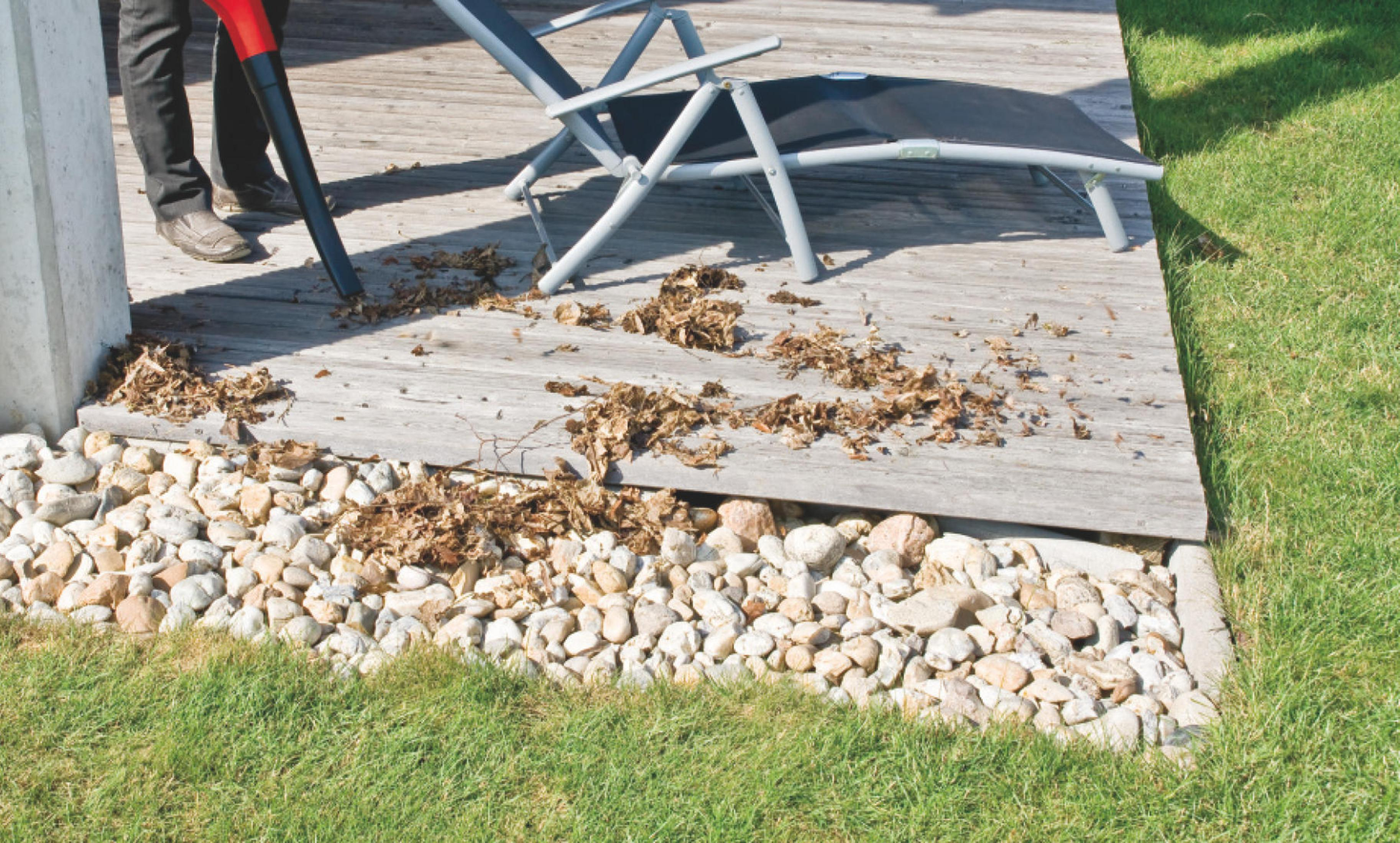 Ein Mann benutzt ein Laubgebläse auf einer Holzterrasse. Es liegen verstreute Blätter und Steine im Bild.