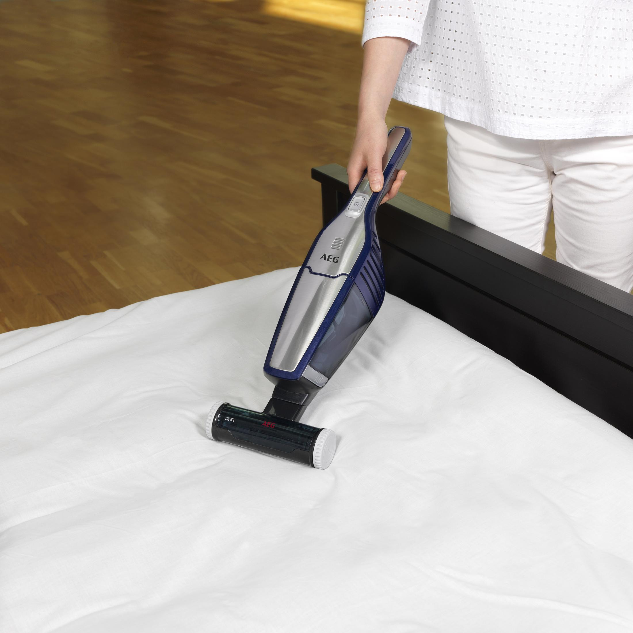 Mujer aspirando una cama con una aspiradora de mano. La cama es blanca y la aspiradora es azul.