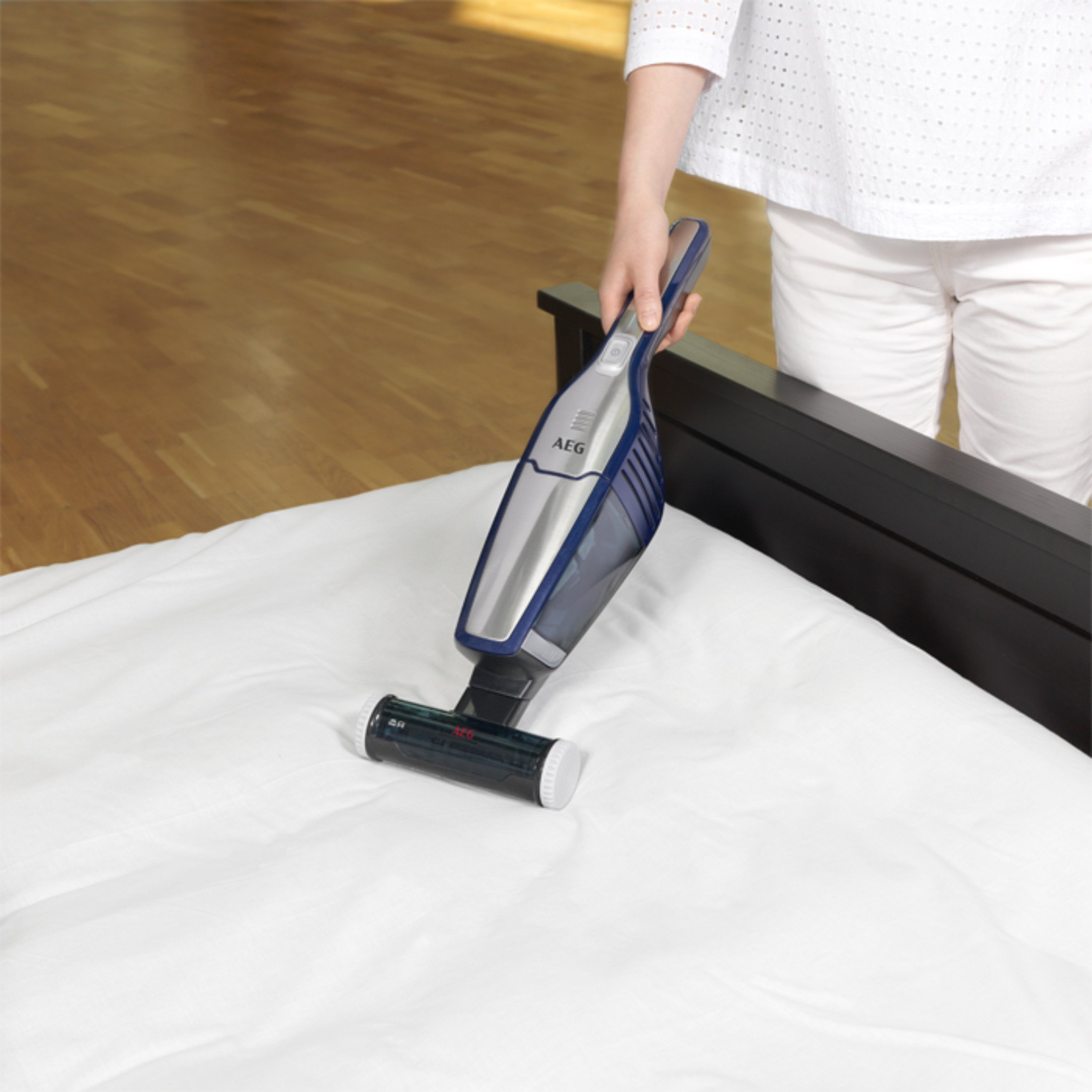Osoba odkurzająca białe łóżko za pomocą niebiesko-srebrnego odkurzacza ręcznego.