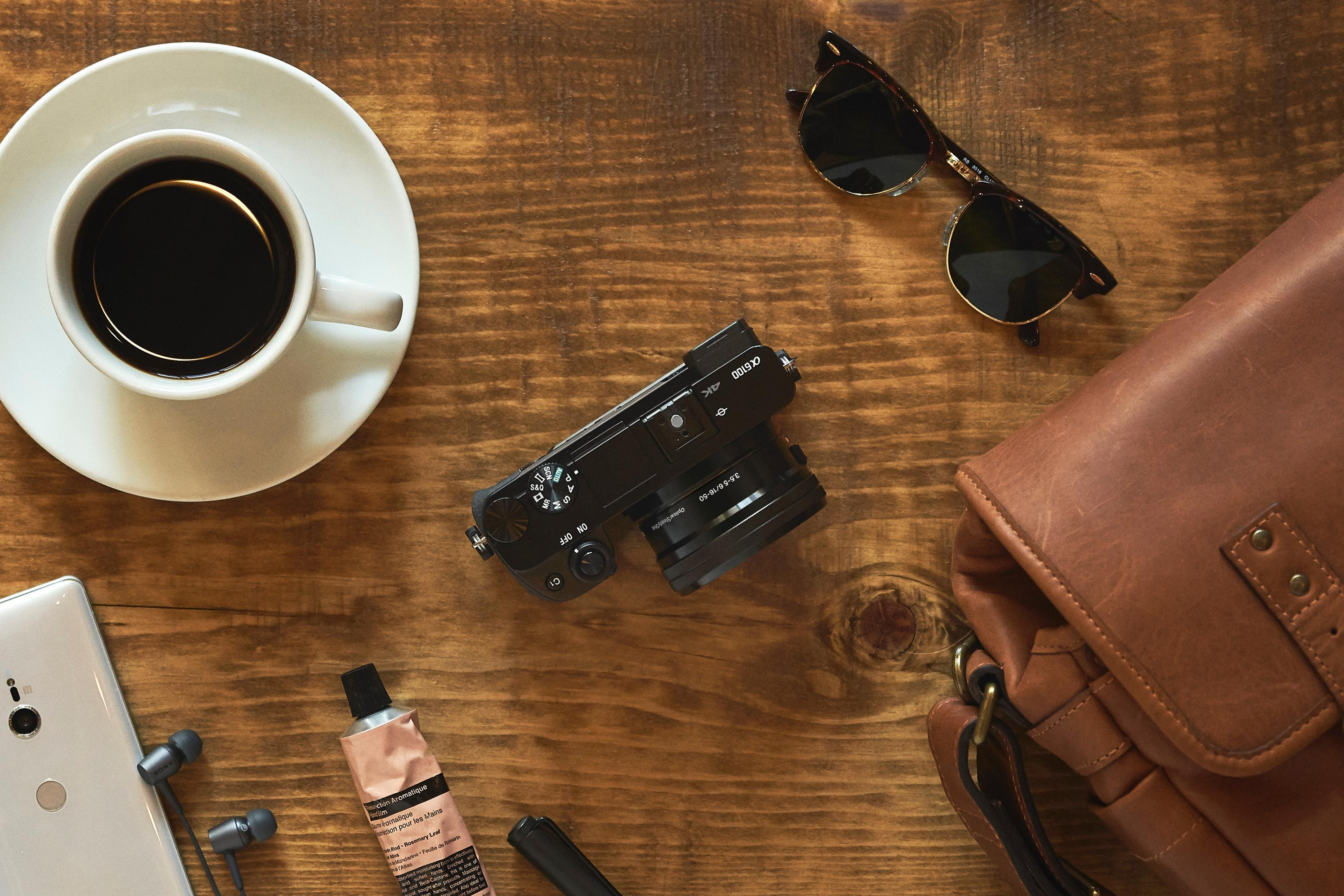 Vista superior de una mesa de madera. Cámara, taza de café, gafas de sol, bolso, teléfono, crema y auriculares.