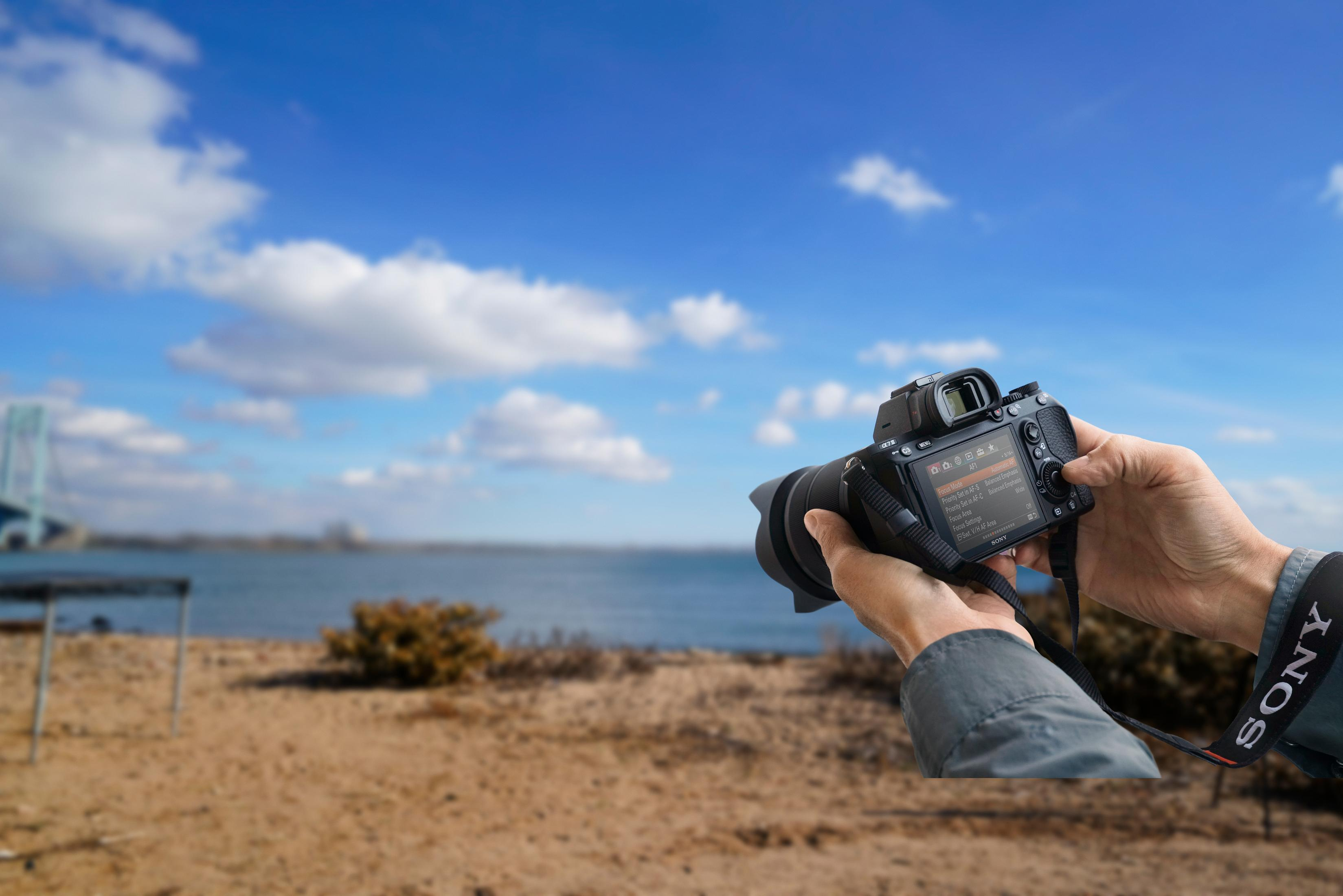 A person holding a camera, taking a photo of a blurred scene with a blue sky and water.
