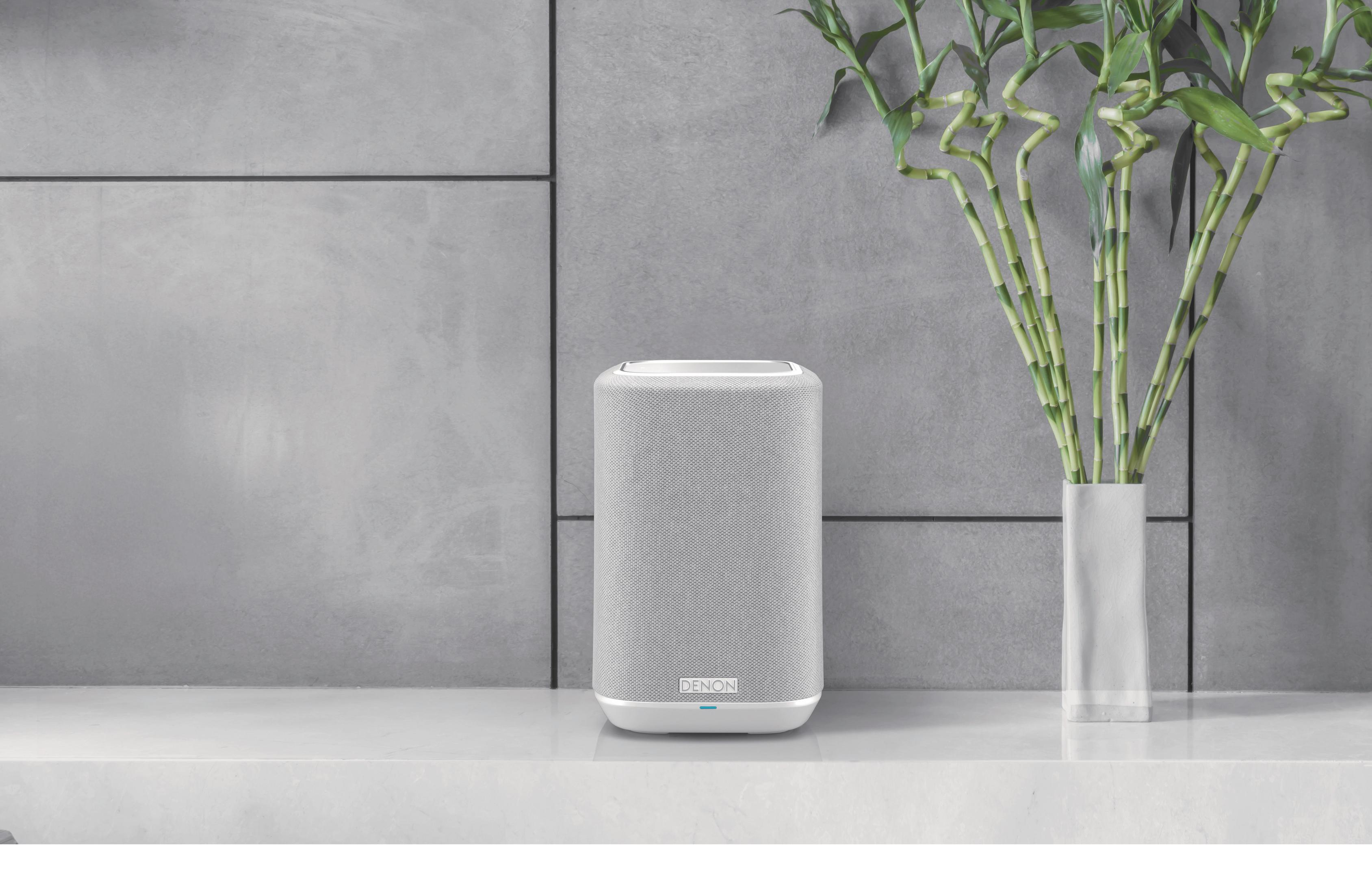 Ein weißer Lautsprecher der Marke Denon auf einer weißen Oberfläche neben einer Vase.