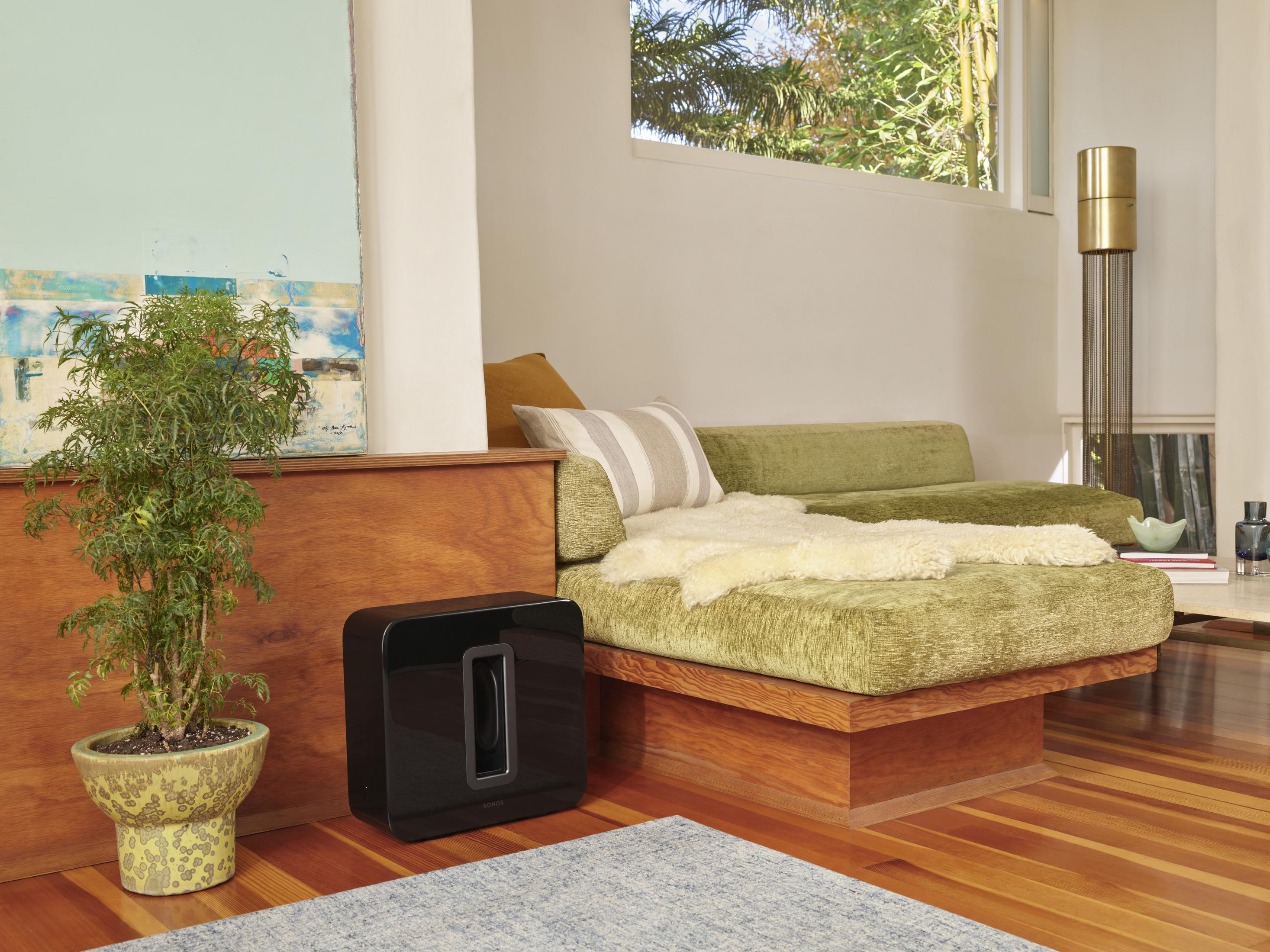 Wohnzimmer mit schwarzem Subwoofer neben einem grünen Sofa und einer Pflanze.