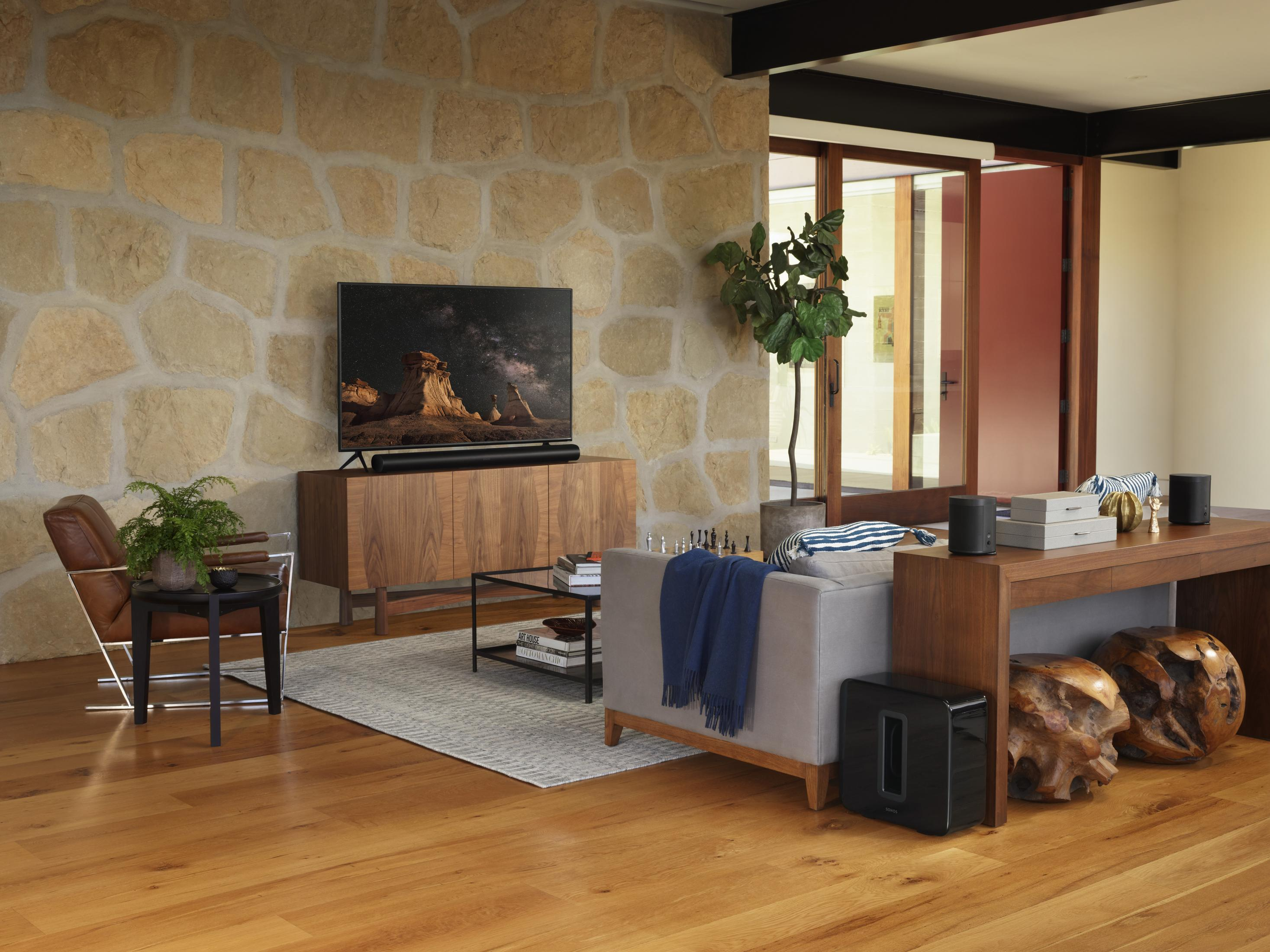 Wohnzimmer mit Fernseher, Lautsprecher und anderen Geräten. Steinwand, Holzboden und Pflanzen sind vorhanden.