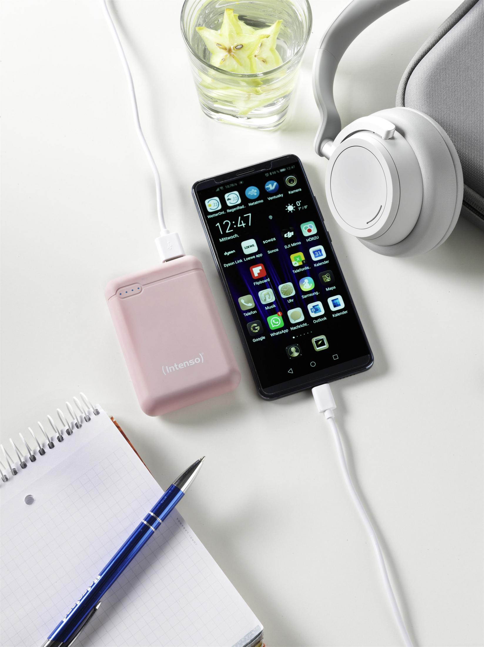 Smartphone wird mit einer pinken Powerbank geladen. Kopfhörer, Stift und Notizbuch liegen auf einem weißen Tisch.