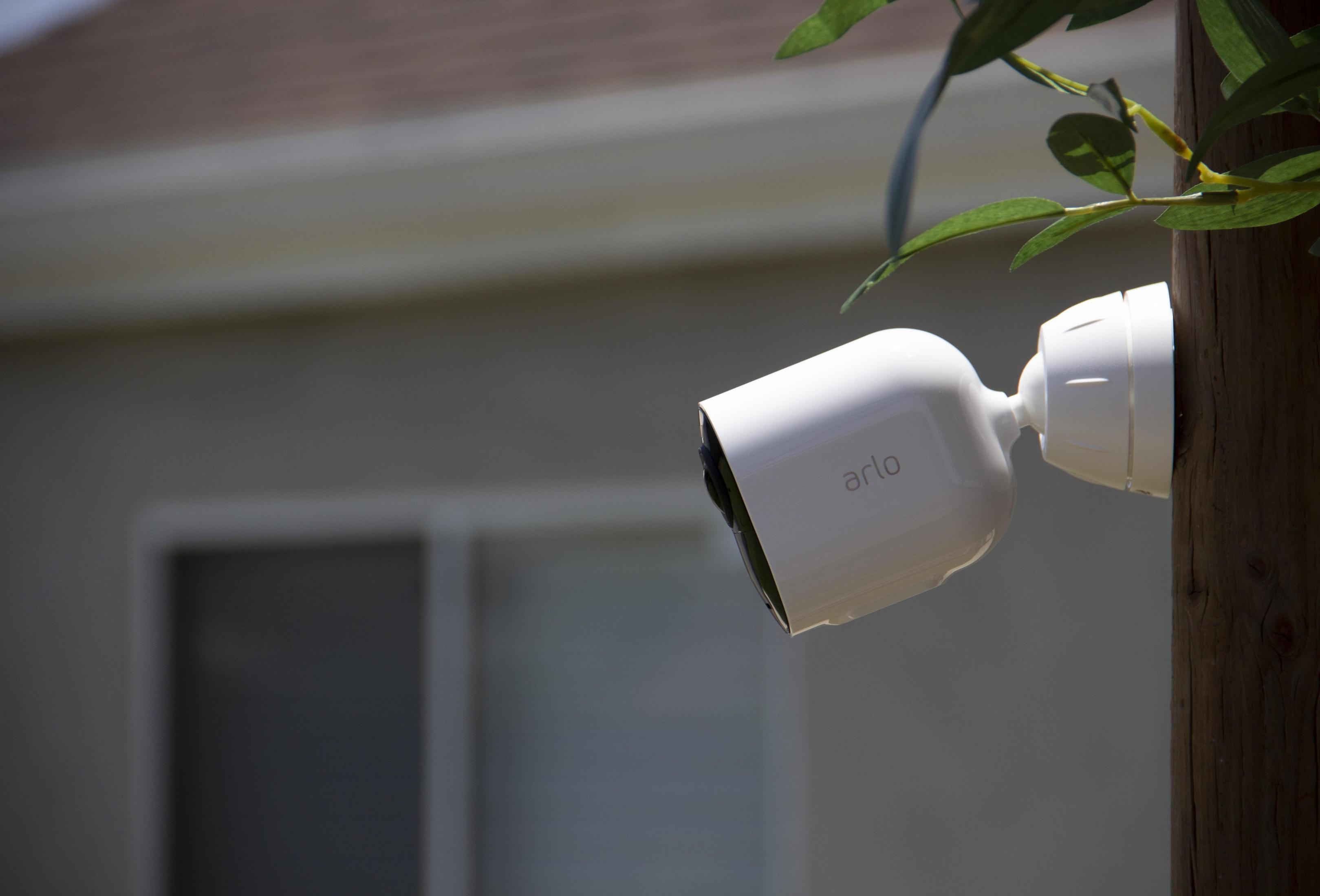 Biała kamera bezpieczeństwa Arlo zamontowana na drewnianym słupie, z rozmytym domem w tle.