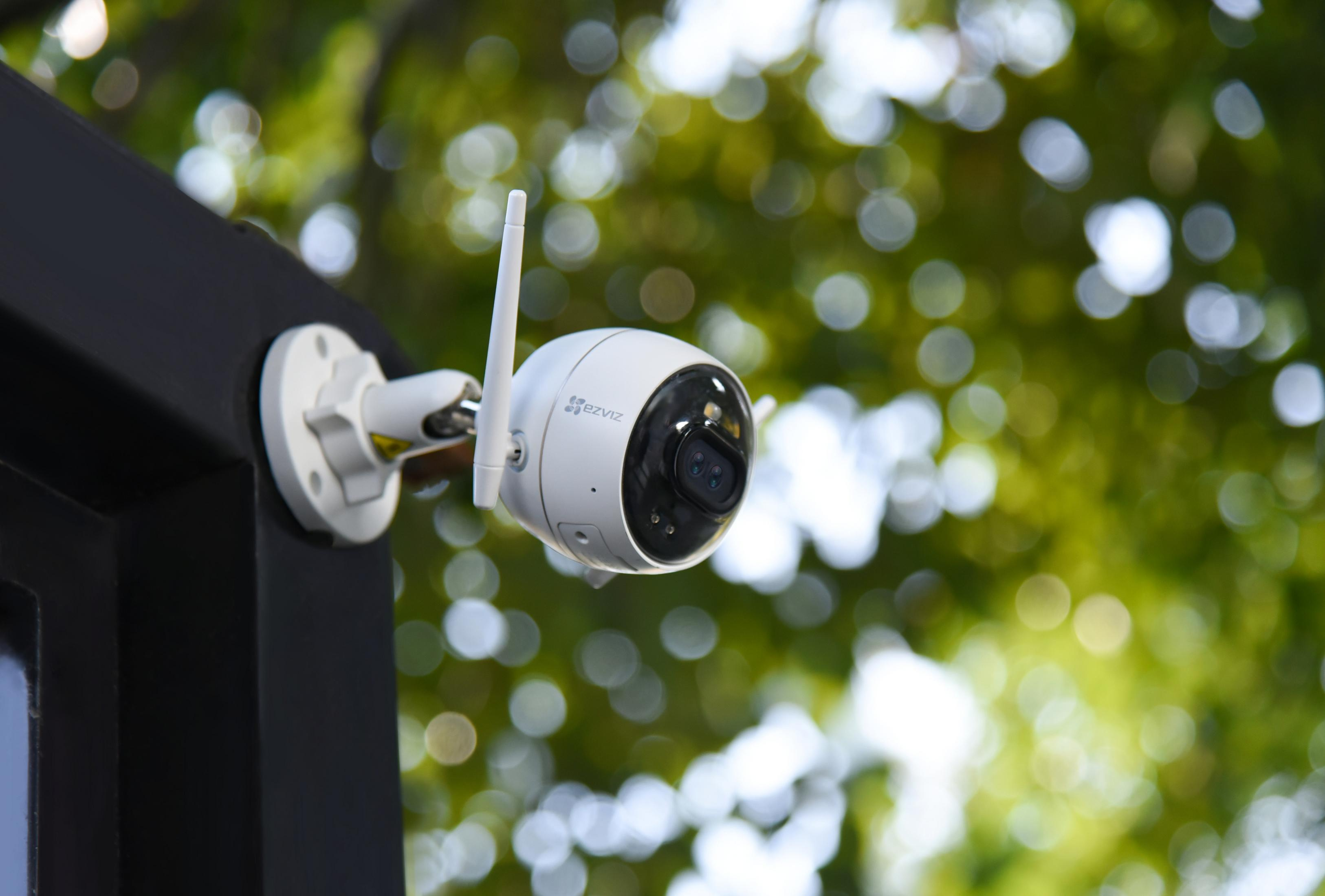 Una telecamera di sicurezza esterna bianca montata su una struttura scura, con alberi verdi sullo sfondo.