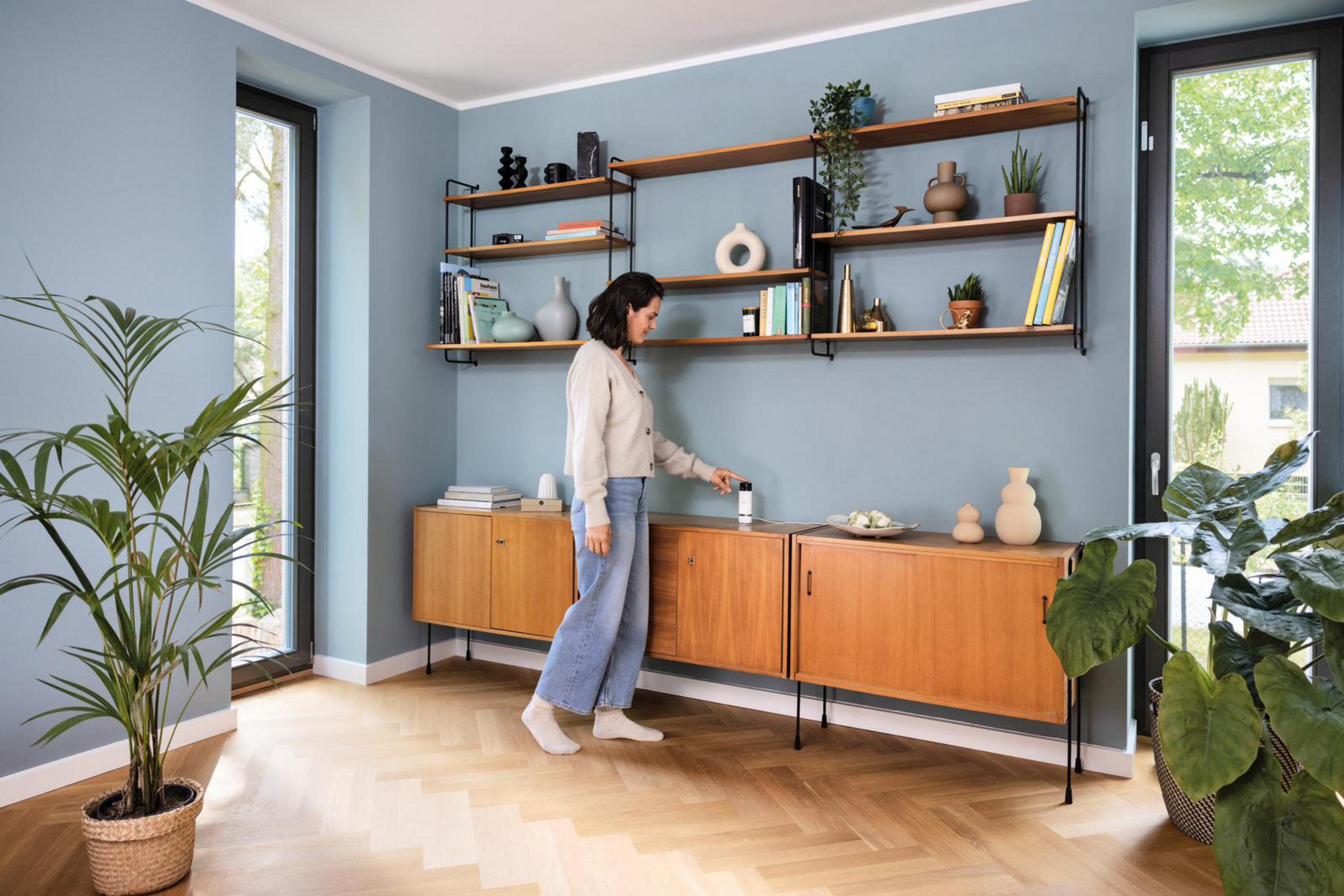 Eine Frau in einem Wohnzimmer berührt ein Smart Home-Gerät auf einem Schrank.