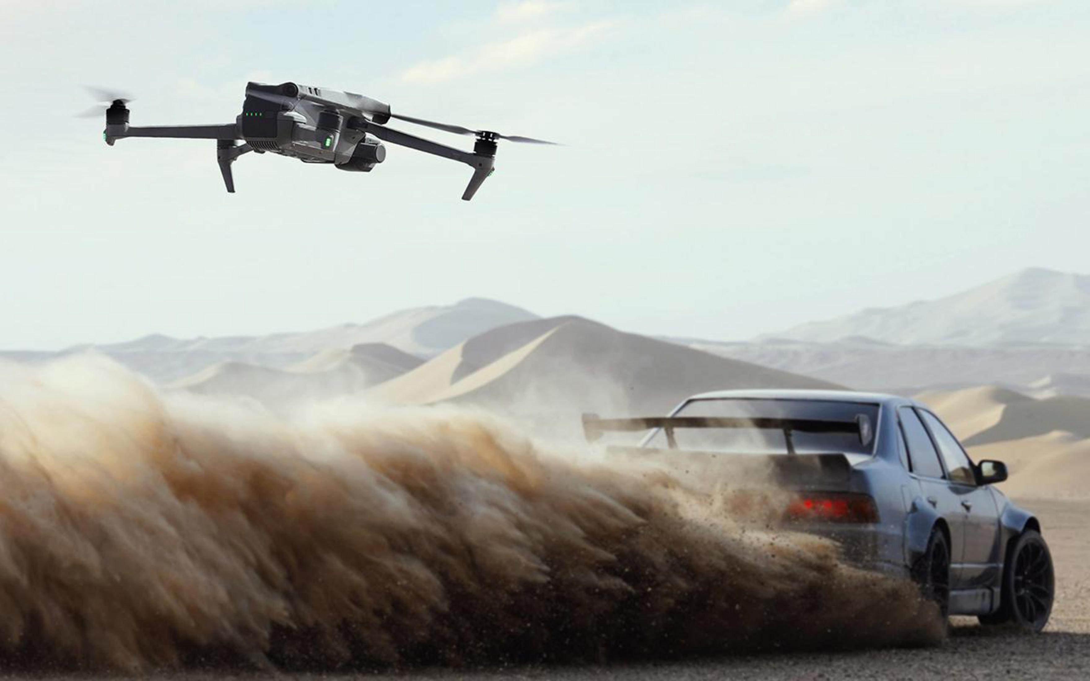 Eine Drohne fliegt am Himmel über einem Auto, das in einer Wüstenkulisse eine Staubwolke aufwirbelt.