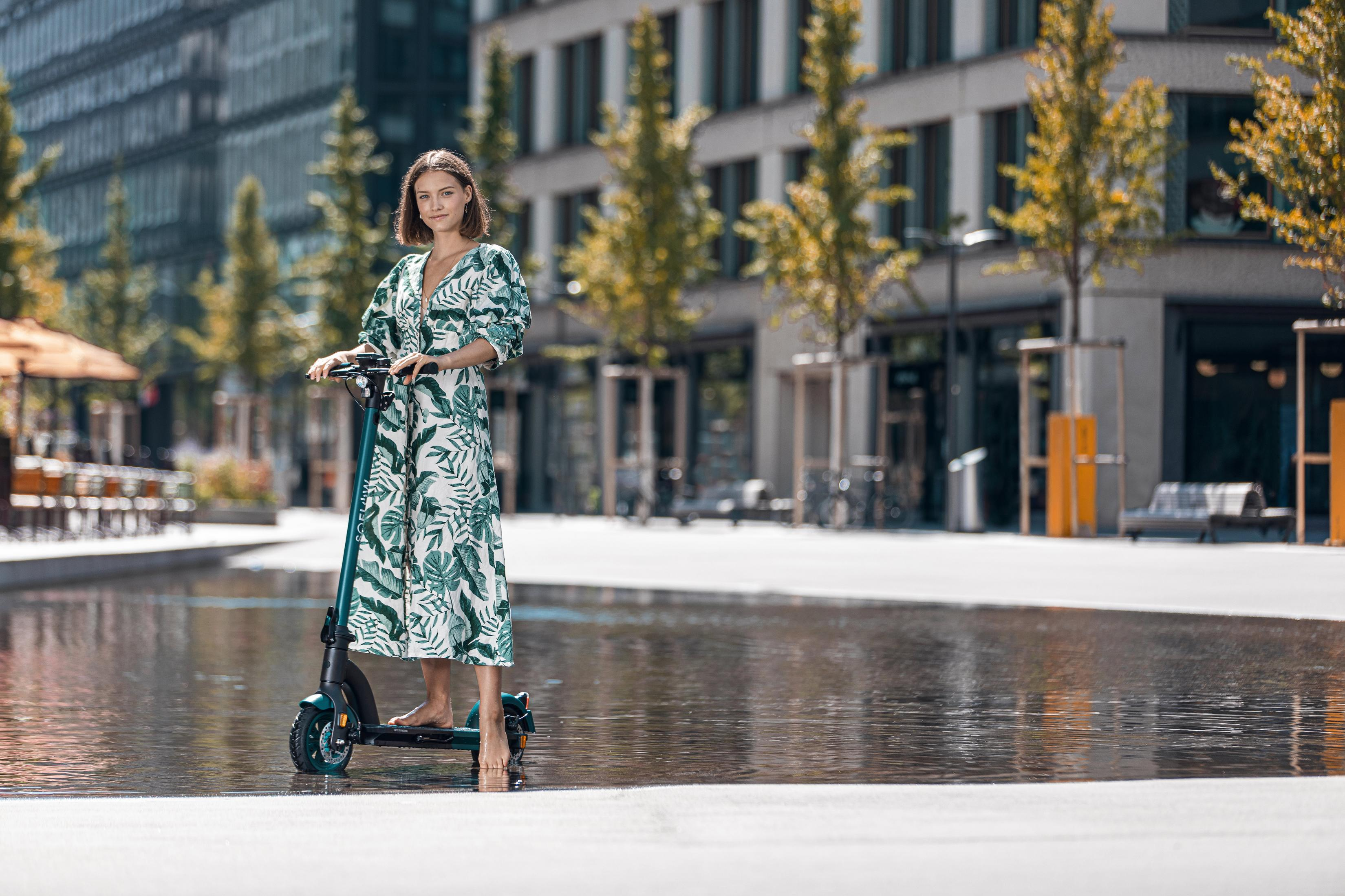Frau fährt einen Elektroroller. Sie trägt ein Kleid und steht auf einem Roller.