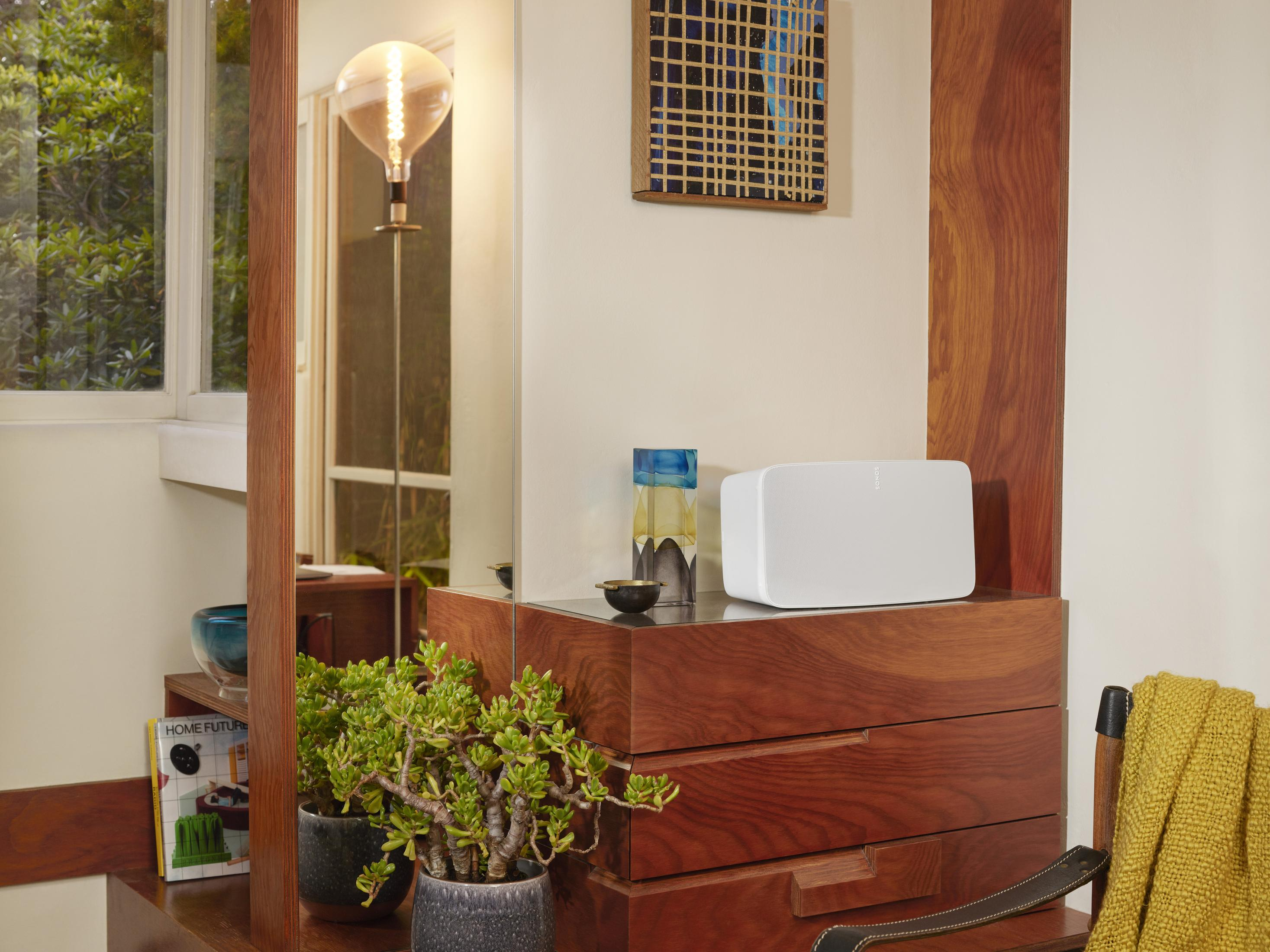 Salon z białym głośnikiem na drewnianej szafce. Widoczne lustro i roślina.