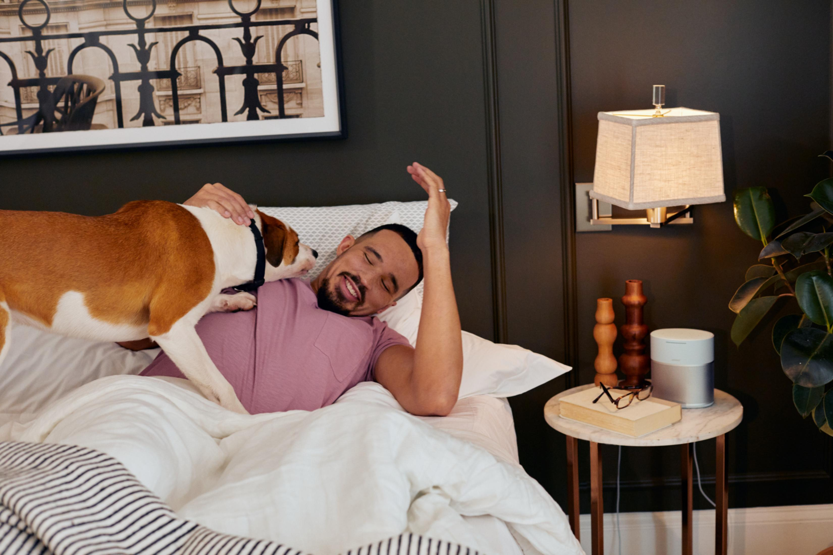 Mann liegt im Bett und streichelt einen Hund. Schlafzimmer mit Lampe und Tisch.