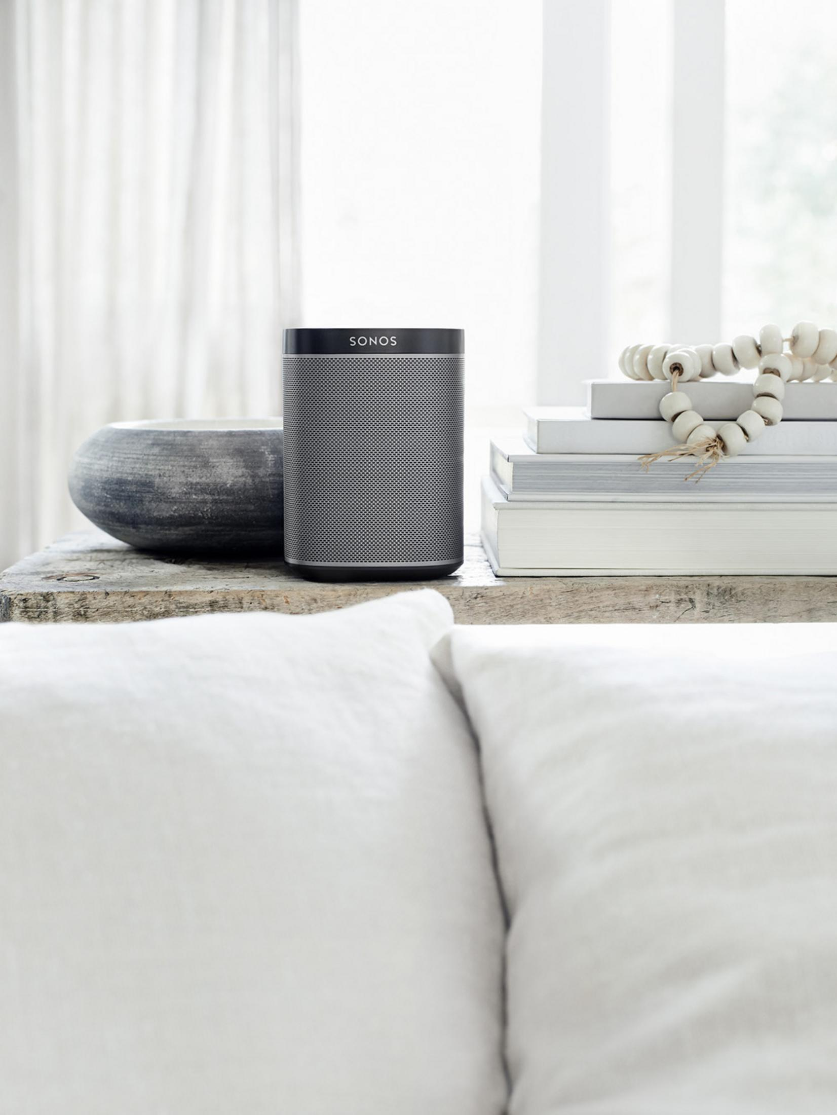 Grauer Sonos-Lautsprecher auf einem Holztisch, mit Büchern und Deko. Weiße Kissen im Vordergrund.