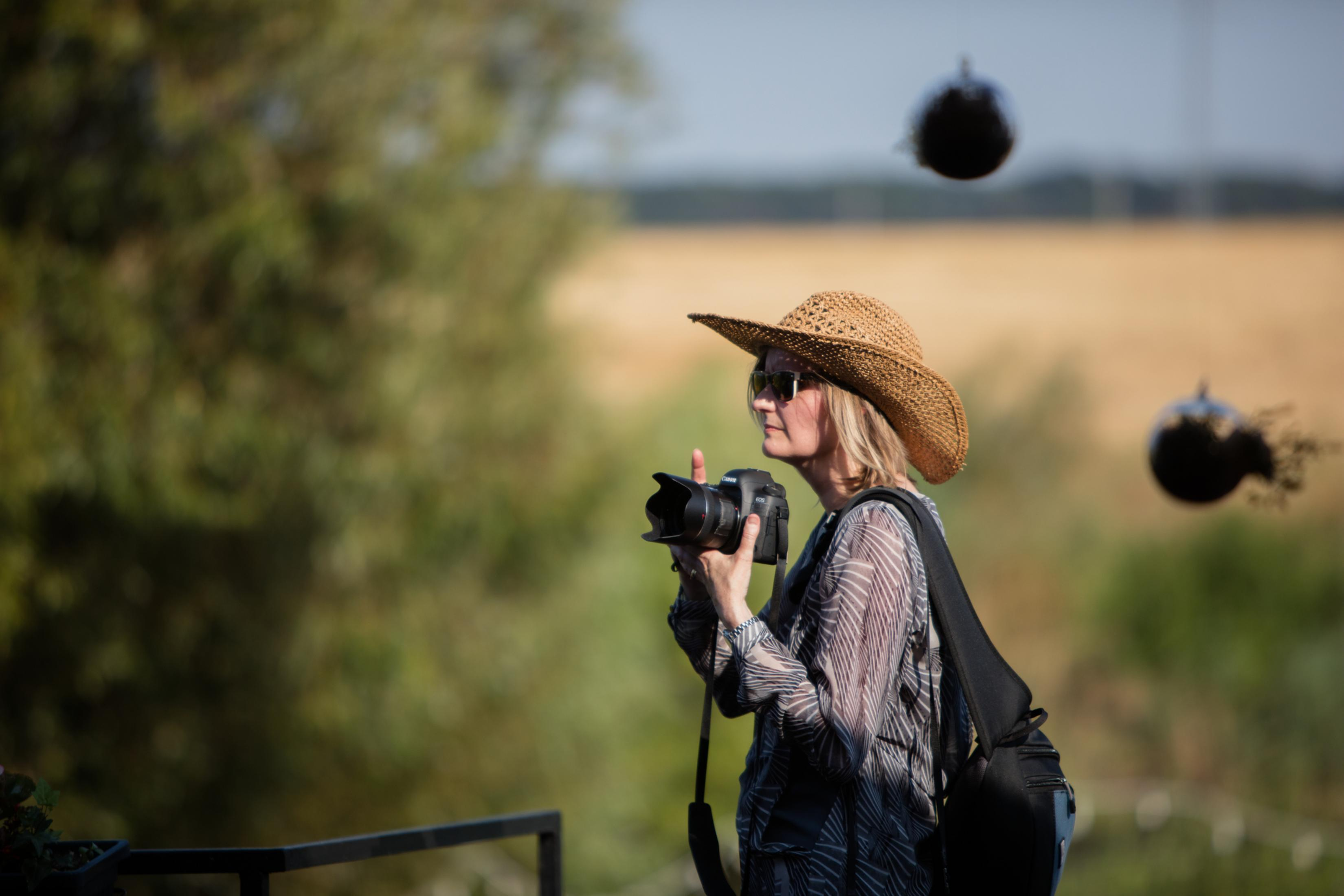 Mujer con sombrero tomando fotos al aire libre con una cámara, fondo borroso con árboles y cielo.
