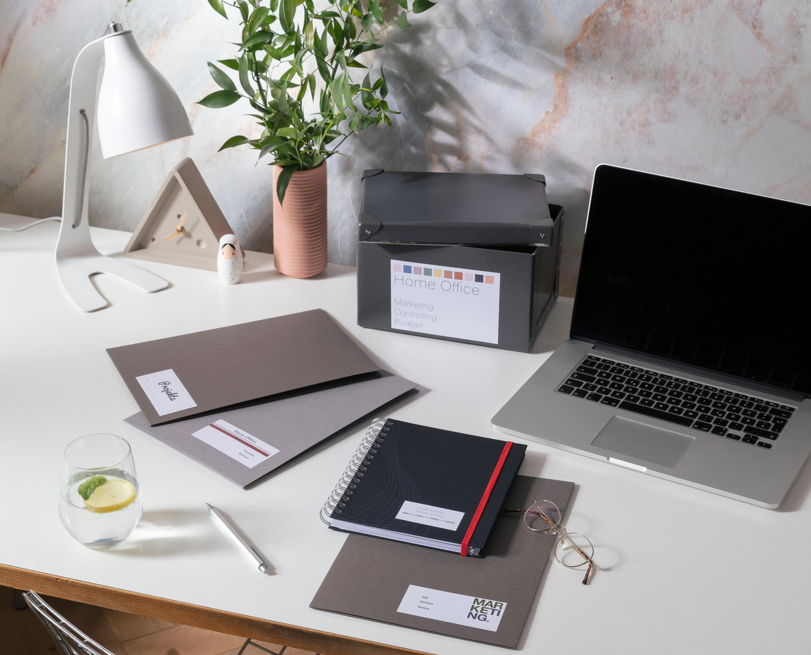 Schreibtisch mit Laptop, Dokumenten, Lampe und Pflanze. Ein Glas Wasser mit Zitrone links.