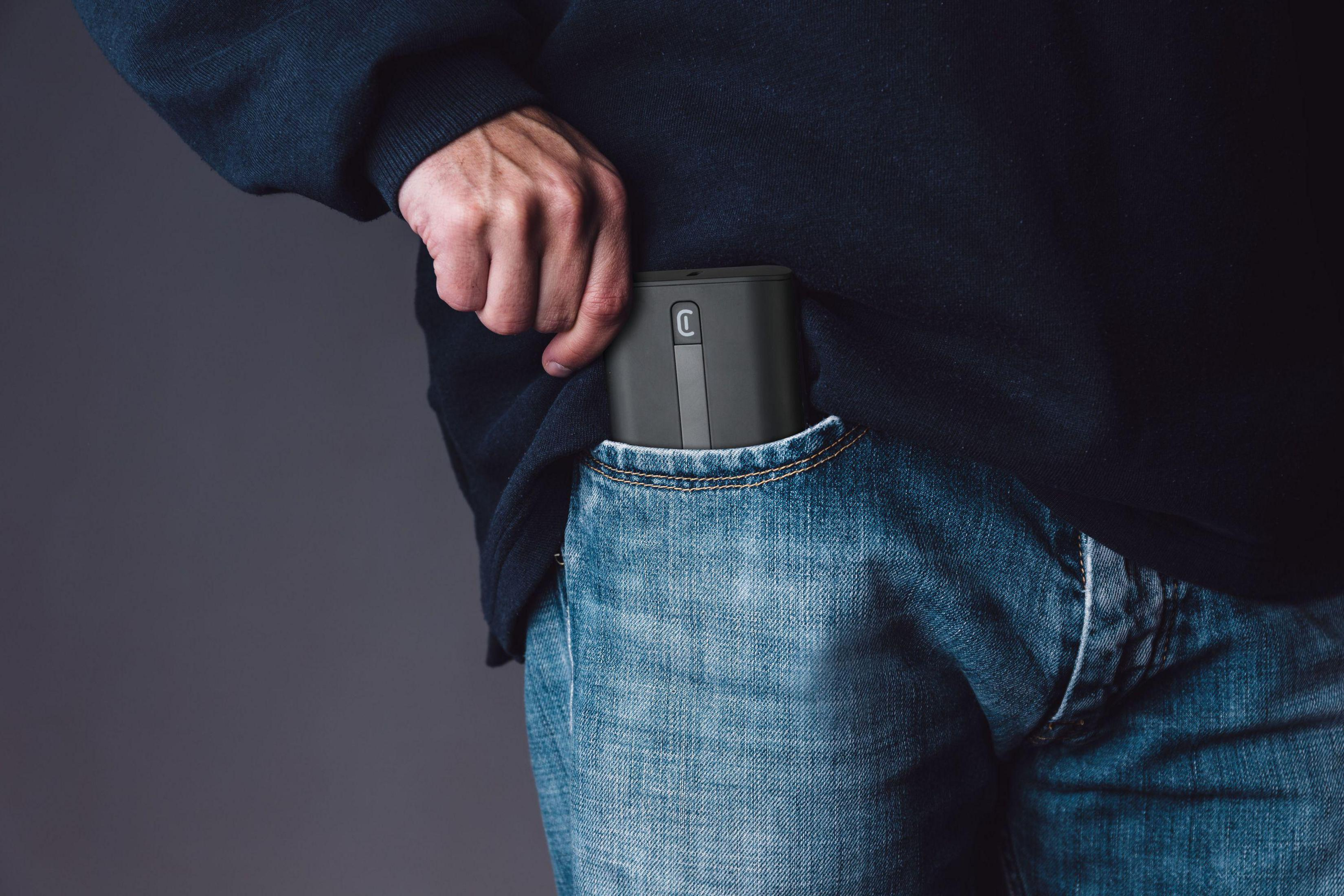 Person in Jeans steckt eine schwarze Powerbank in die Hosentasche. Grauer Hintergrund.