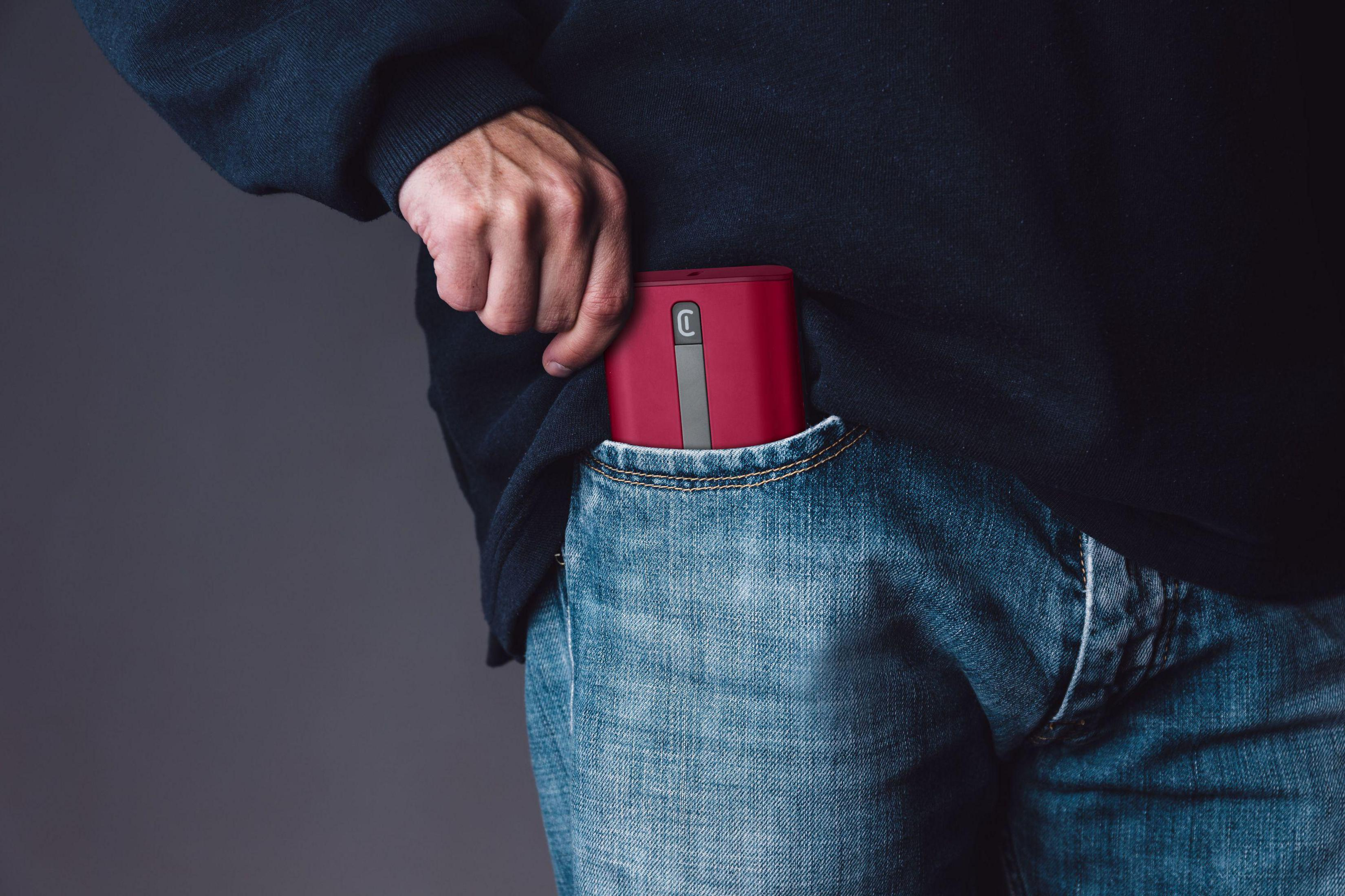 Die Hand einer Person greift in eine Jeans-Tasche und hält ein rot-graues Gerät. Das Gerät ist teilweise sichtbar.