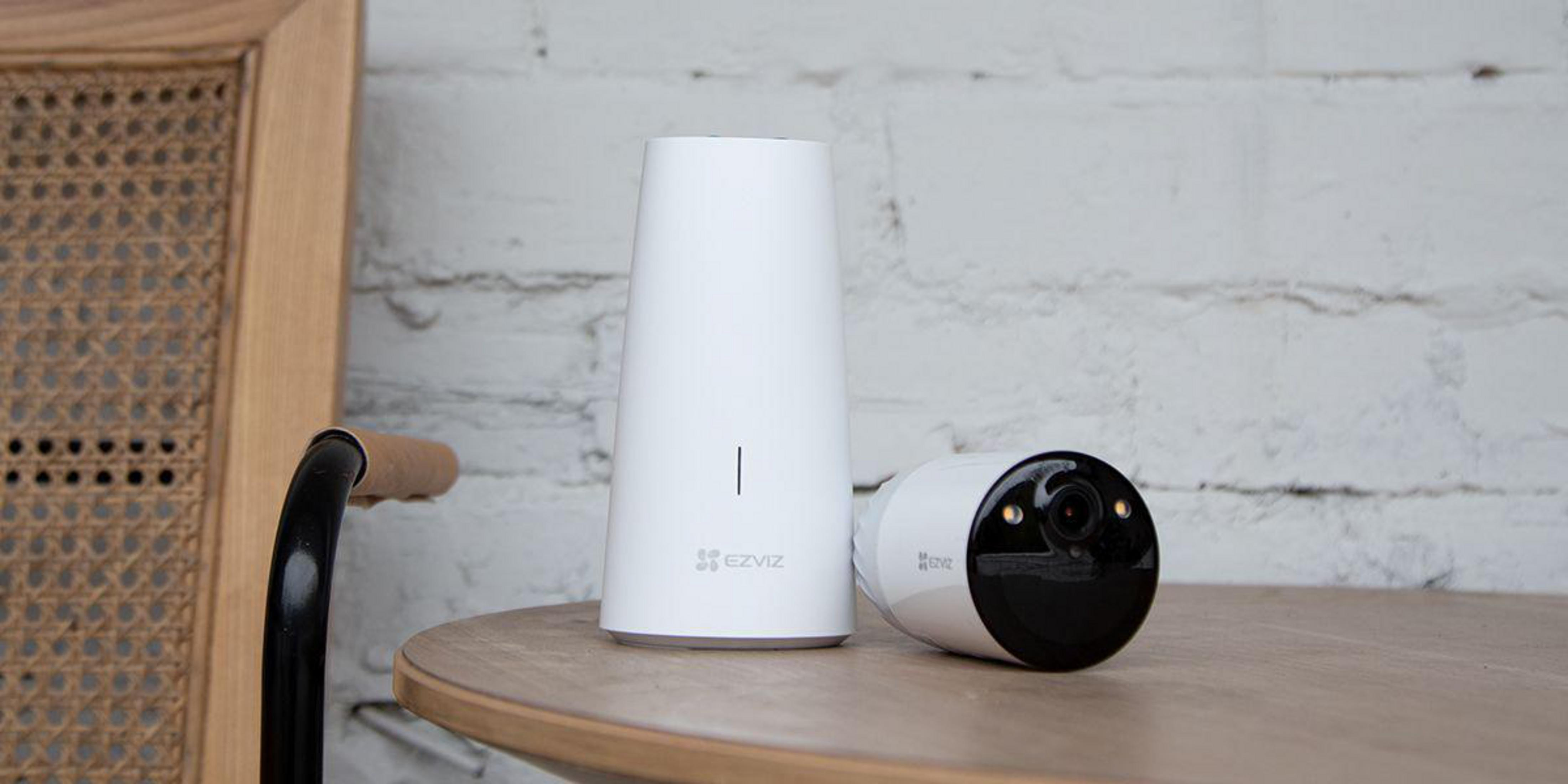 Biała kamera bezpieczeństwa i stacja bazowa na drewnianym stole, tło z ceglanej ściany.