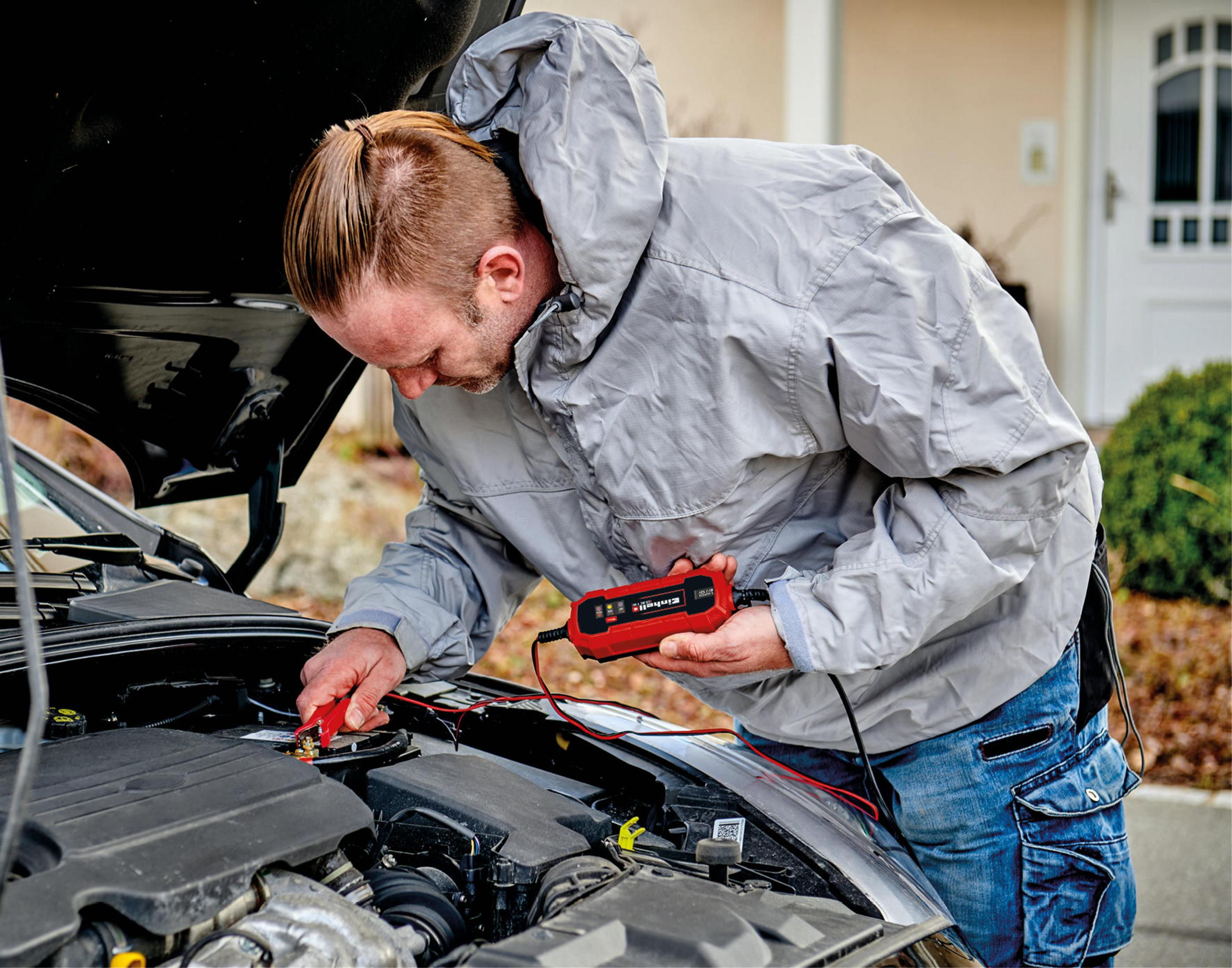 Mann arbeitet an einem Auto und schließt ein rotes Batterieladegerät an die Batterie an.