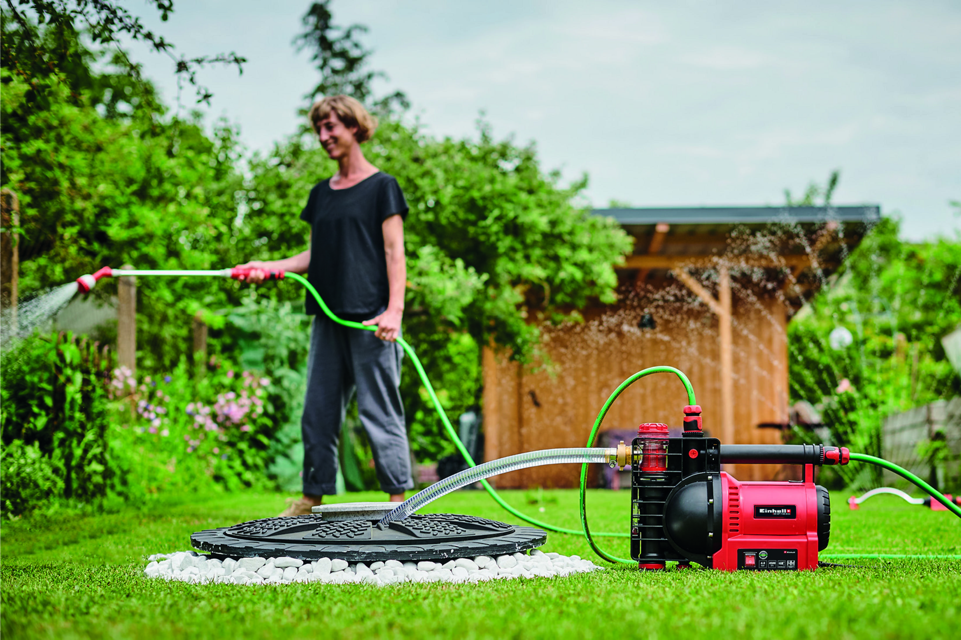 Kobieta podlewa ogród z czerwoną pompą wodną w tle.