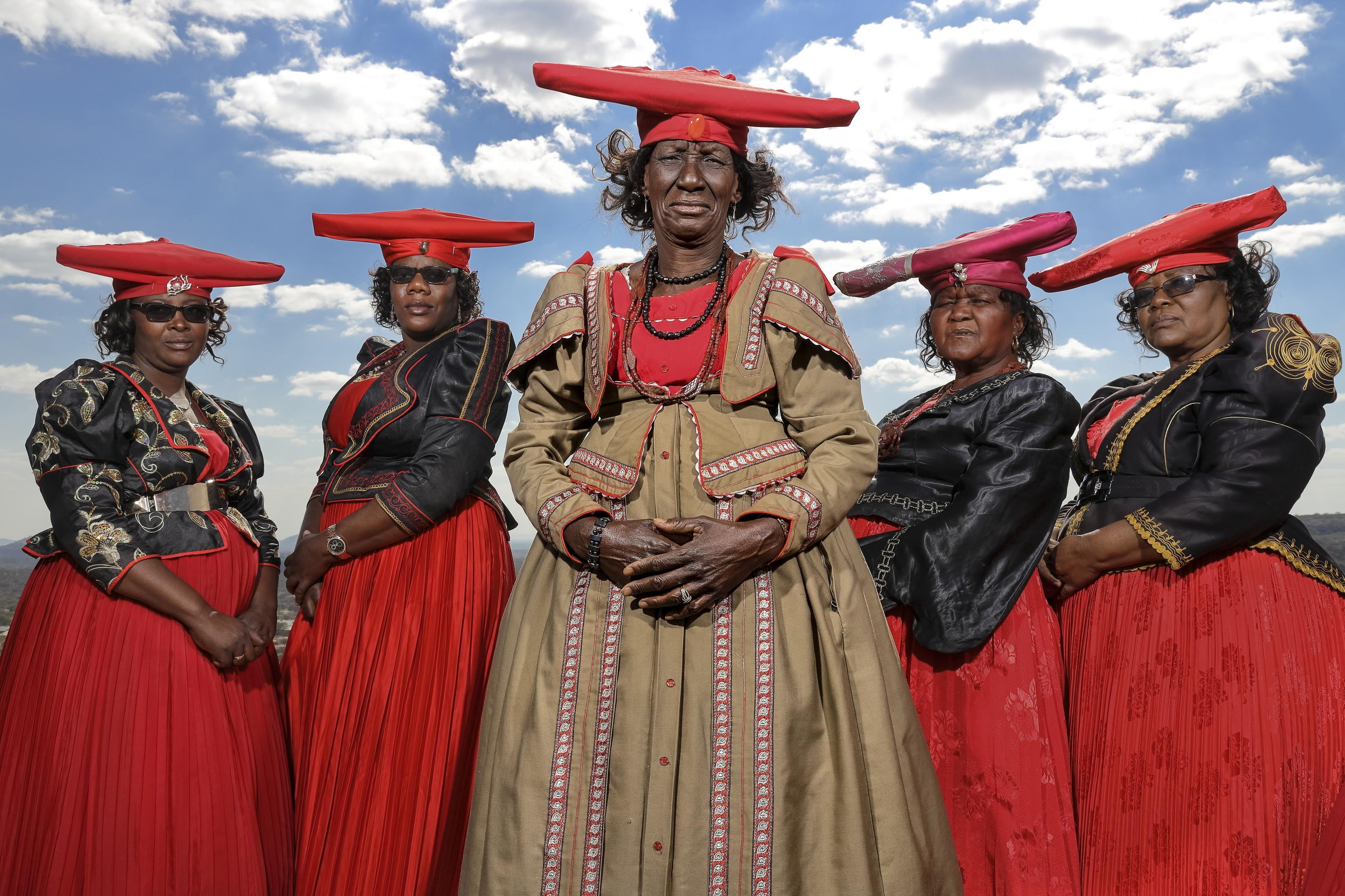 Cinco mujeres con vestimenta tradicional herero, de pie juntas, contra cielo azul.
