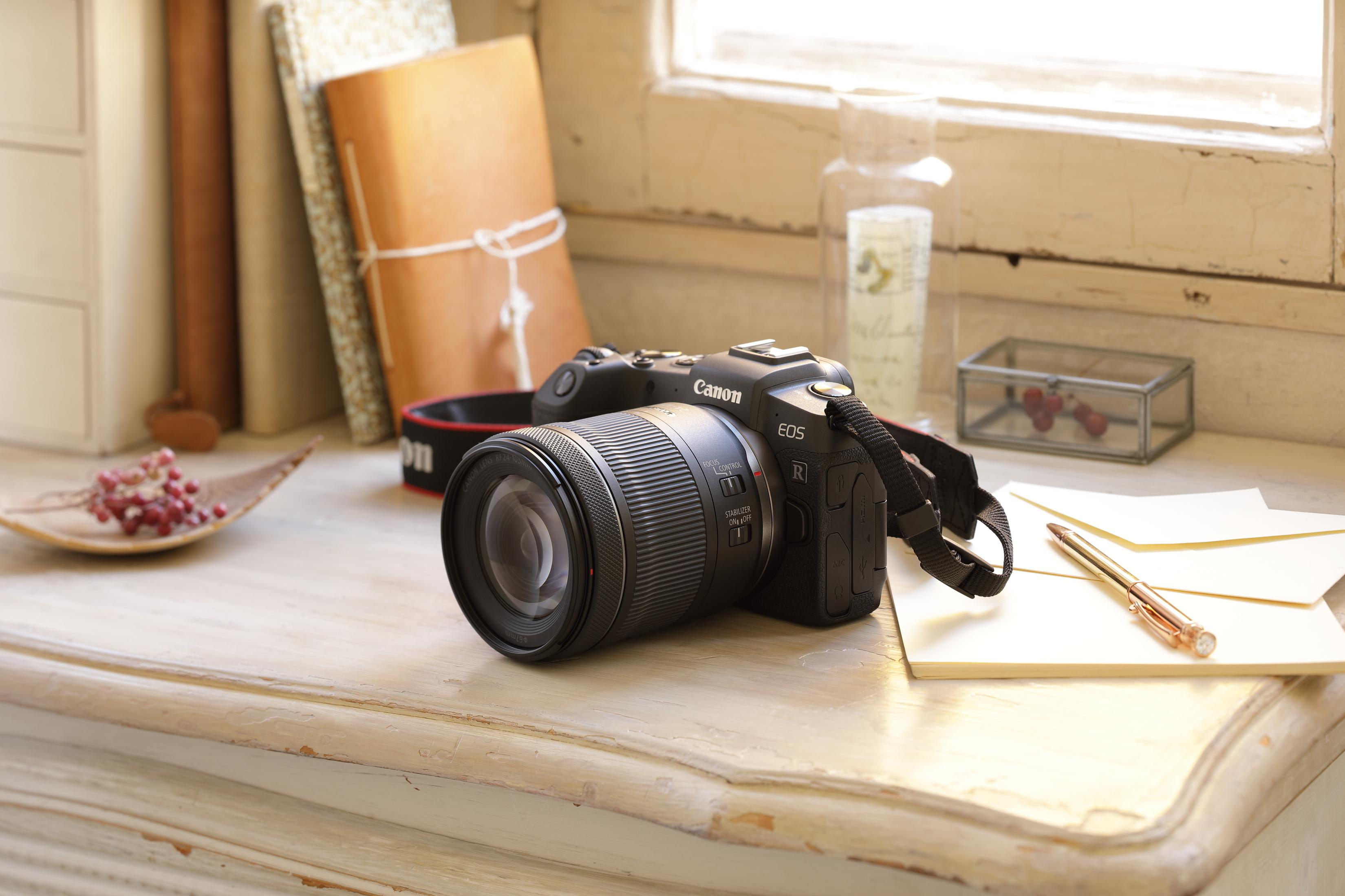 A black camera with a lens and strap, sitting on a white desk. Books and a glass jar are in the background.