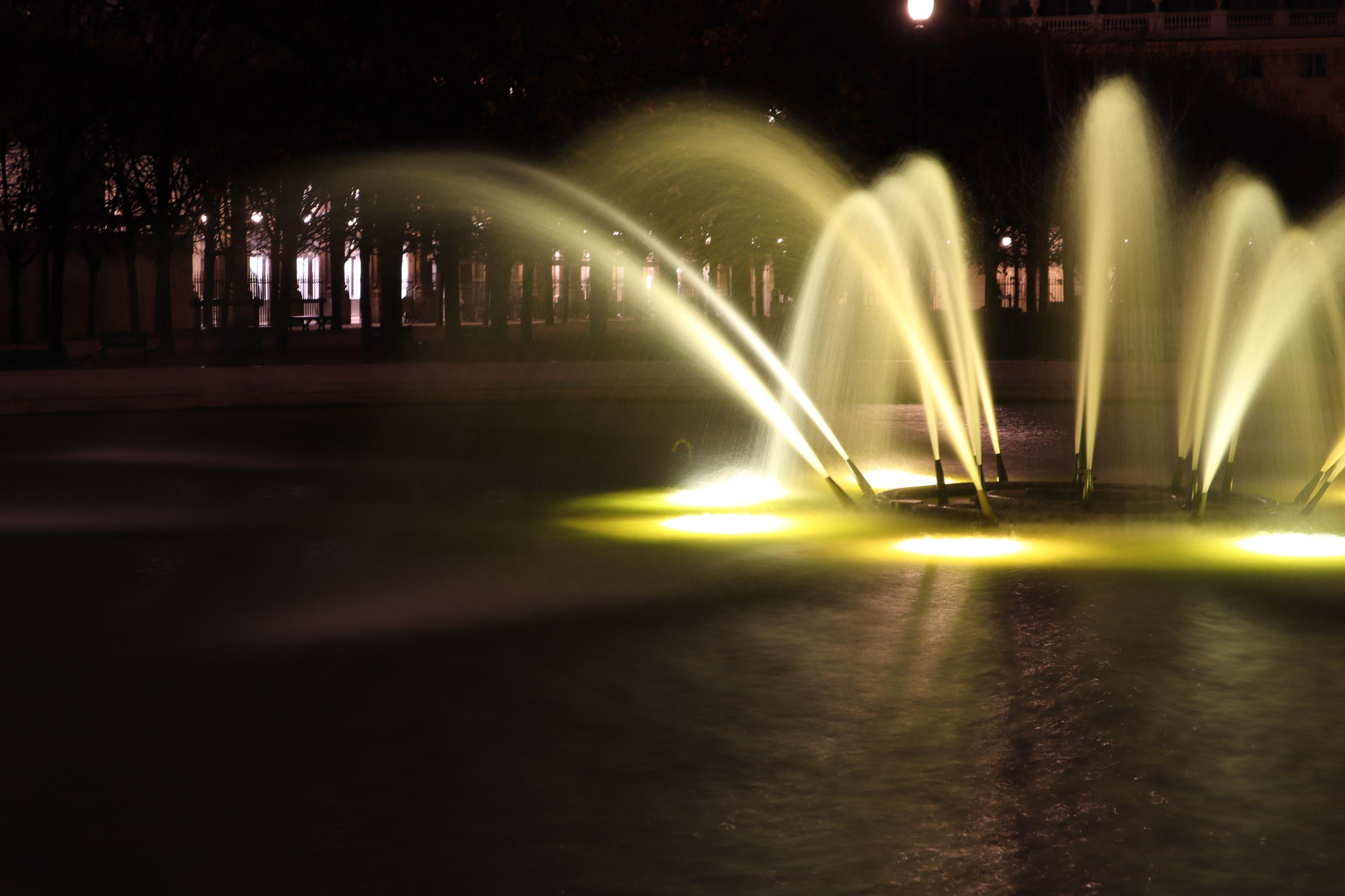 Ein Brunnen bei Nacht mit Bögen aus Wasser, die von gelben Lichtern beleuchtet werden. Dunkler Hintergrund.