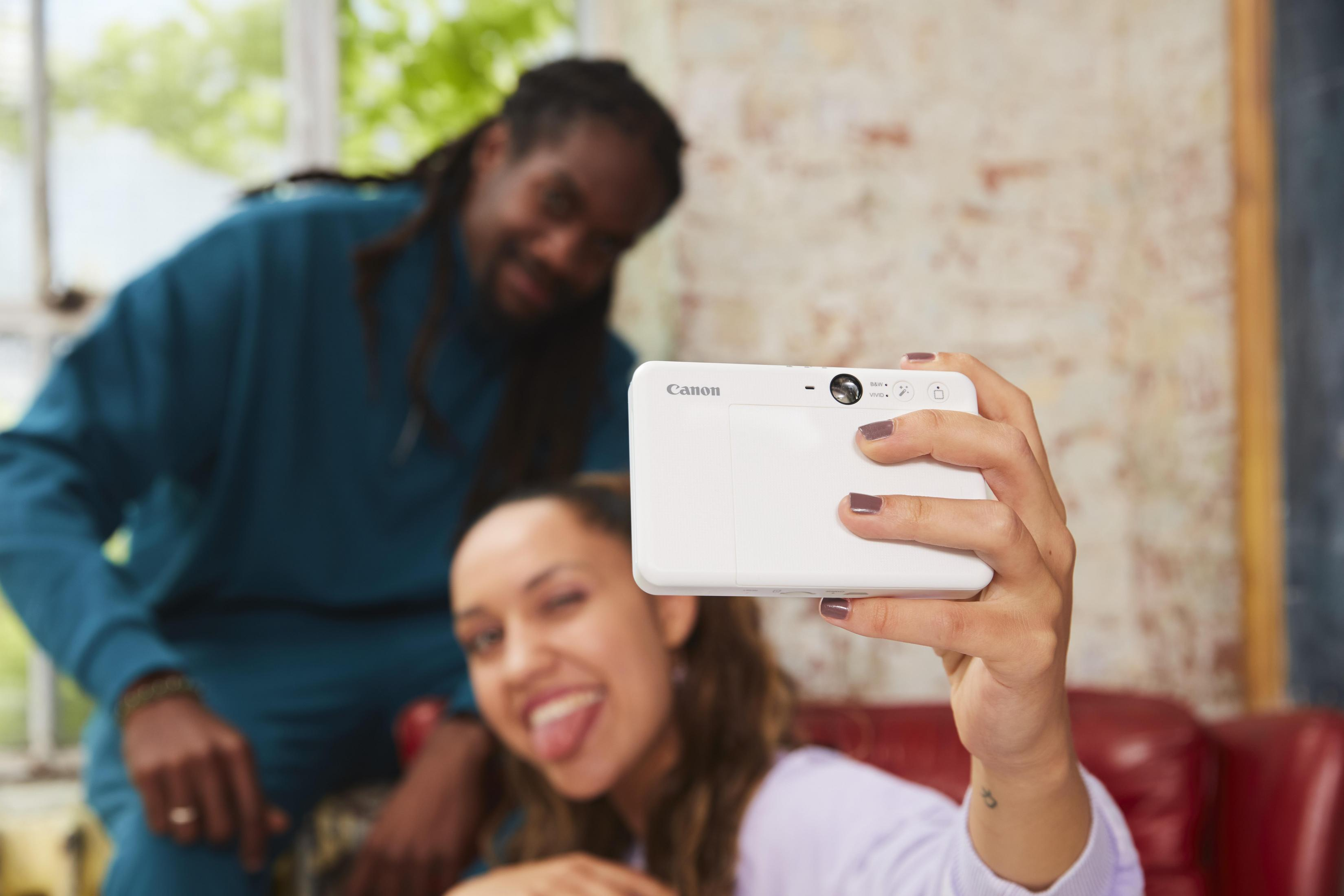 Kobieta robi selfie białą kamerą Canon; mężczyzna uśmiecha się w tle.
