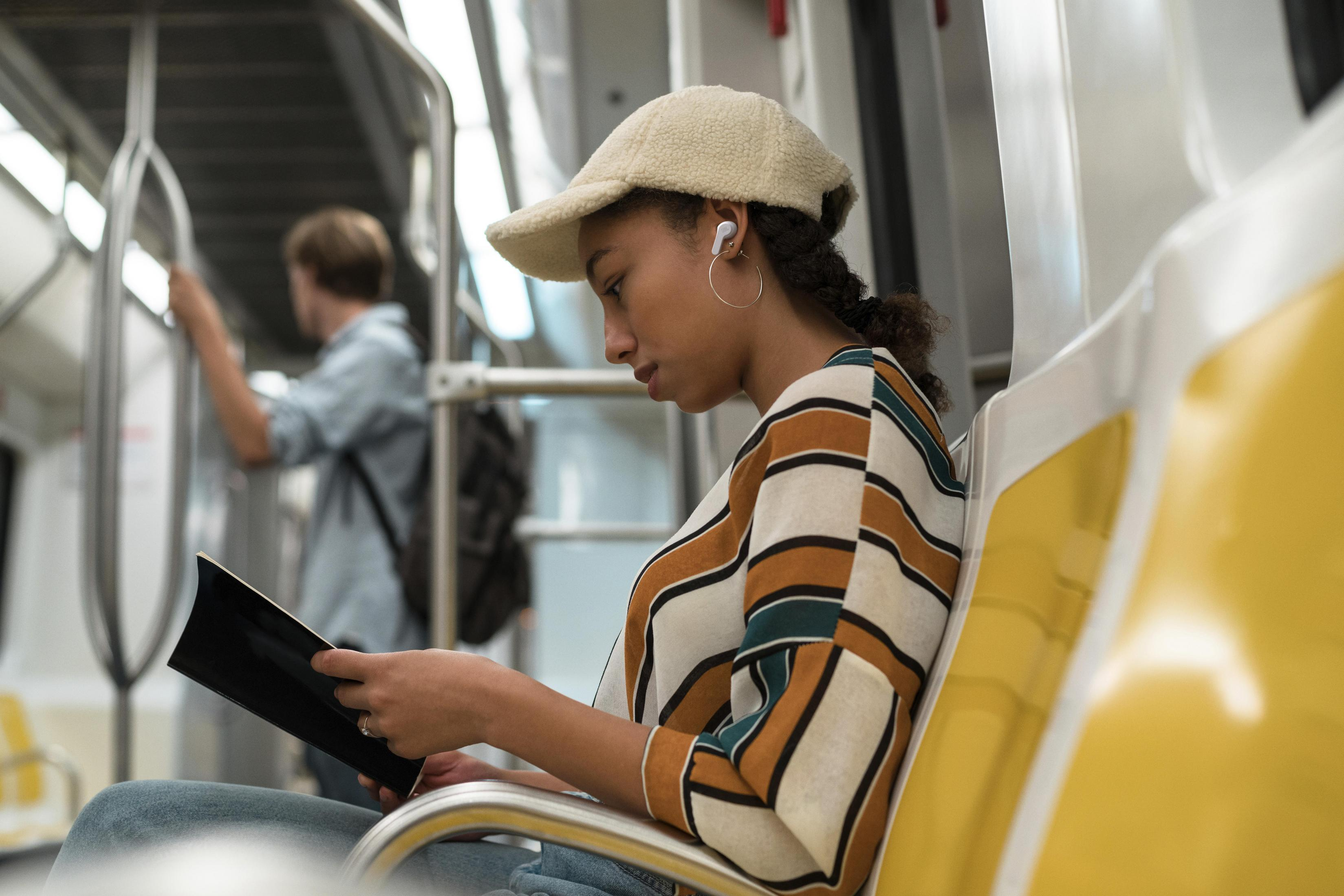 Kobieta w kapeluszu czyta książkę w pociągu metra. Żółte siedzenia i metalowe poręcze.