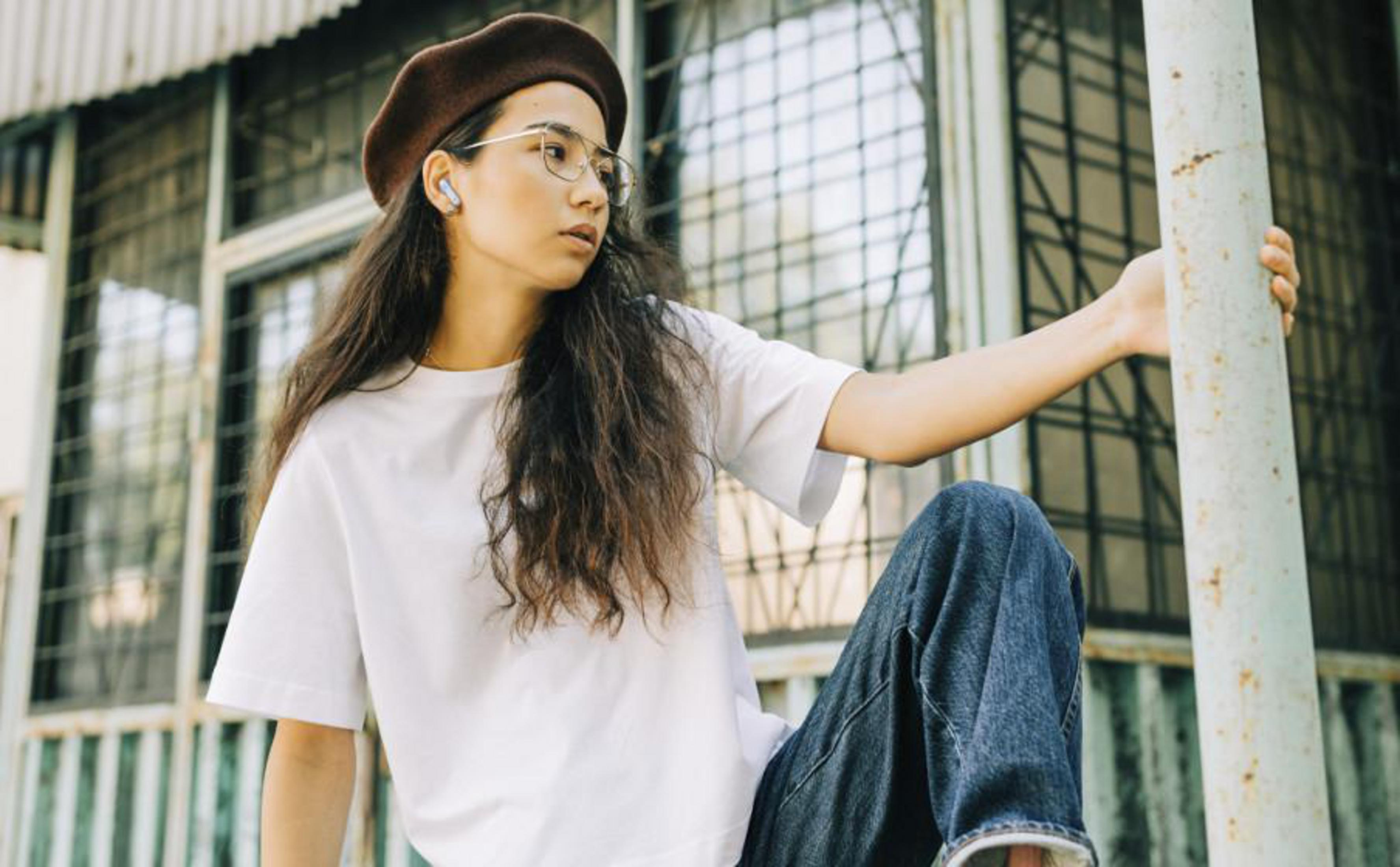 Una donna con occhiali e basco si appoggia a un palo di metallo, indossa una camicia bianca e jeans.