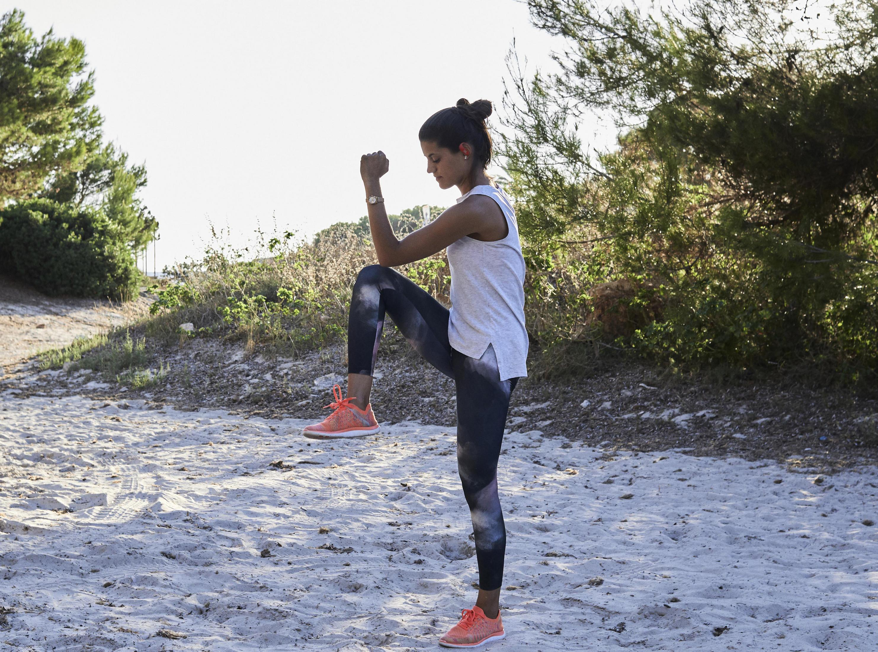 Kobieta w stroju sportowym wykonuje ćwiczenie wysokich kolan na piasku z drzewami w tle.