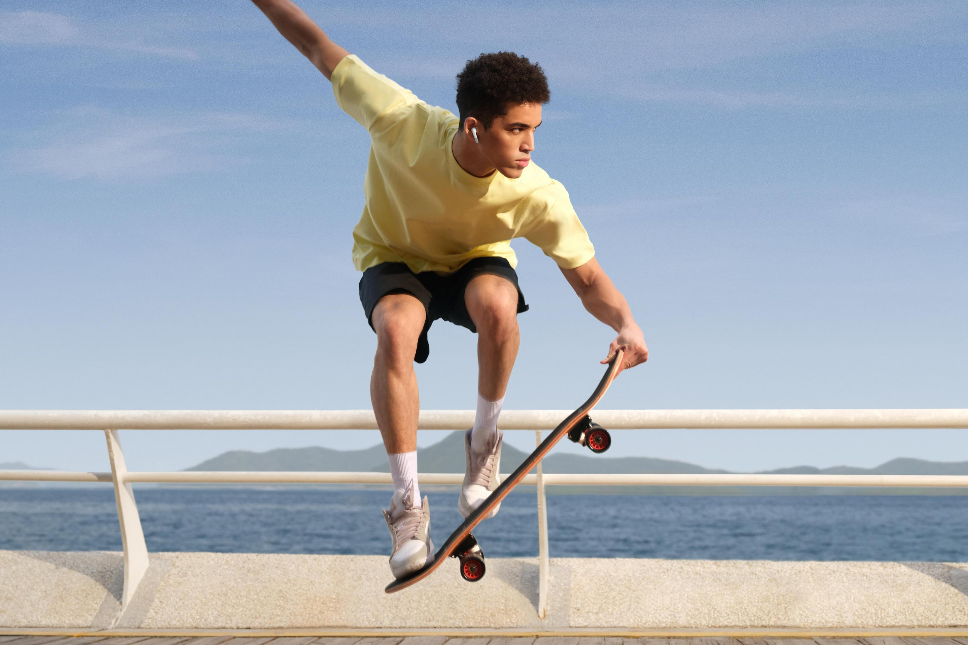 Junger Mann, der auf einem Steg skateboardet. Er ist in der Luft mit dem Board. Blauer Himmel und Wasser im Hintergrund.