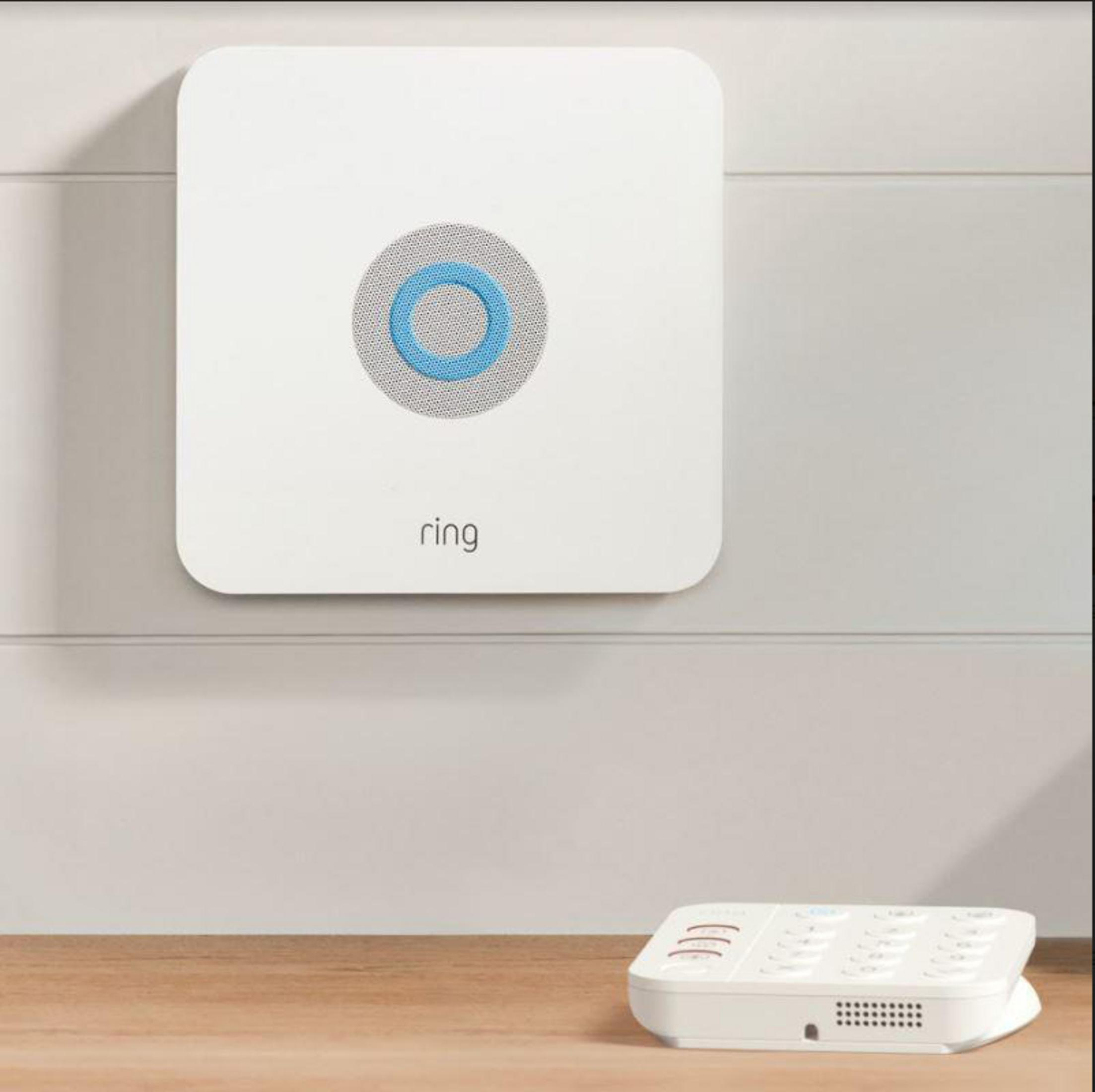 Biały panel alarmowy Ring zamontowany na ścianie i biały telefon na drewnianej powierzchni.