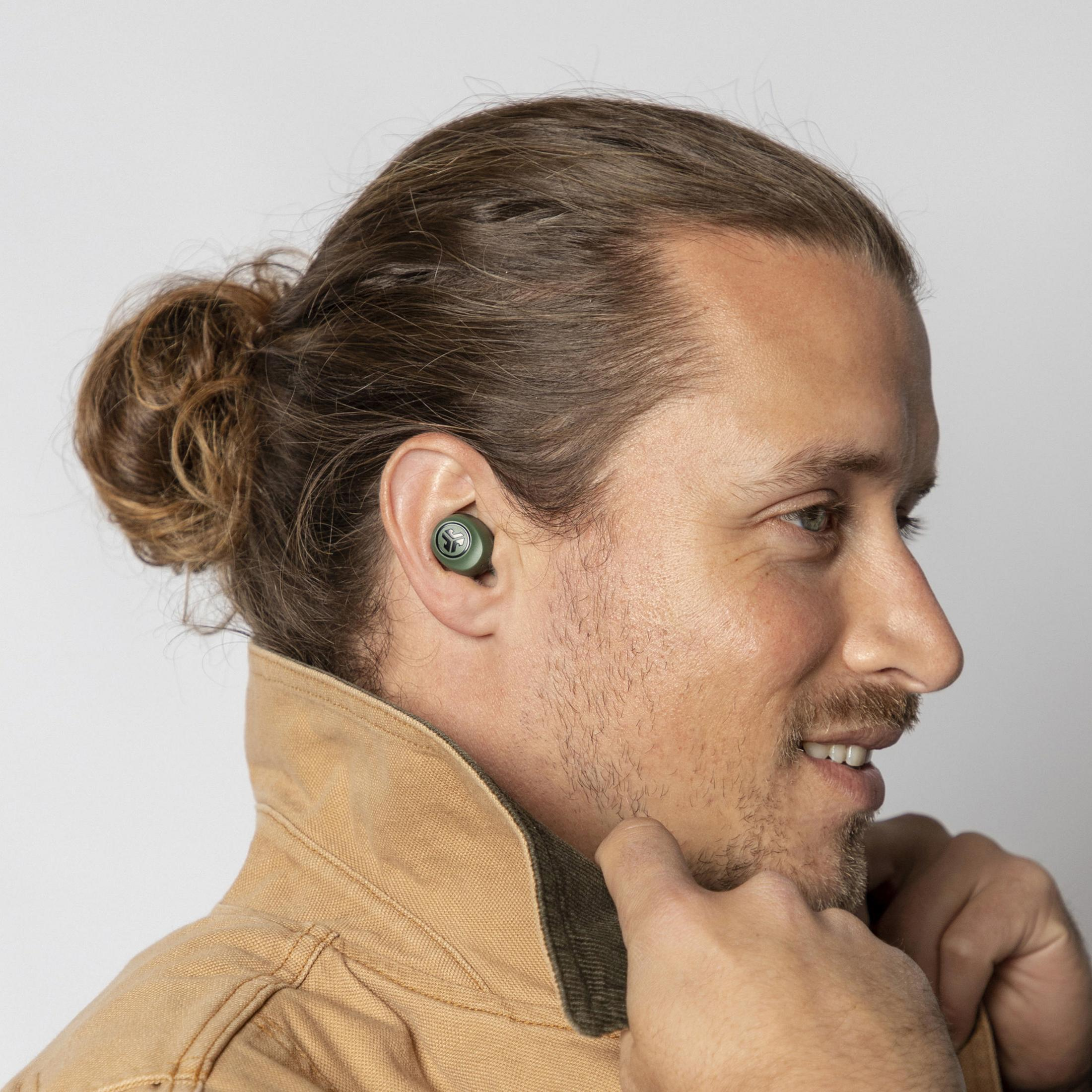 Mężczyzna w słuchawkach, kok. Ciemnozielona słuchawka w uchu, uśmiechnięty, patrzy w prawo.