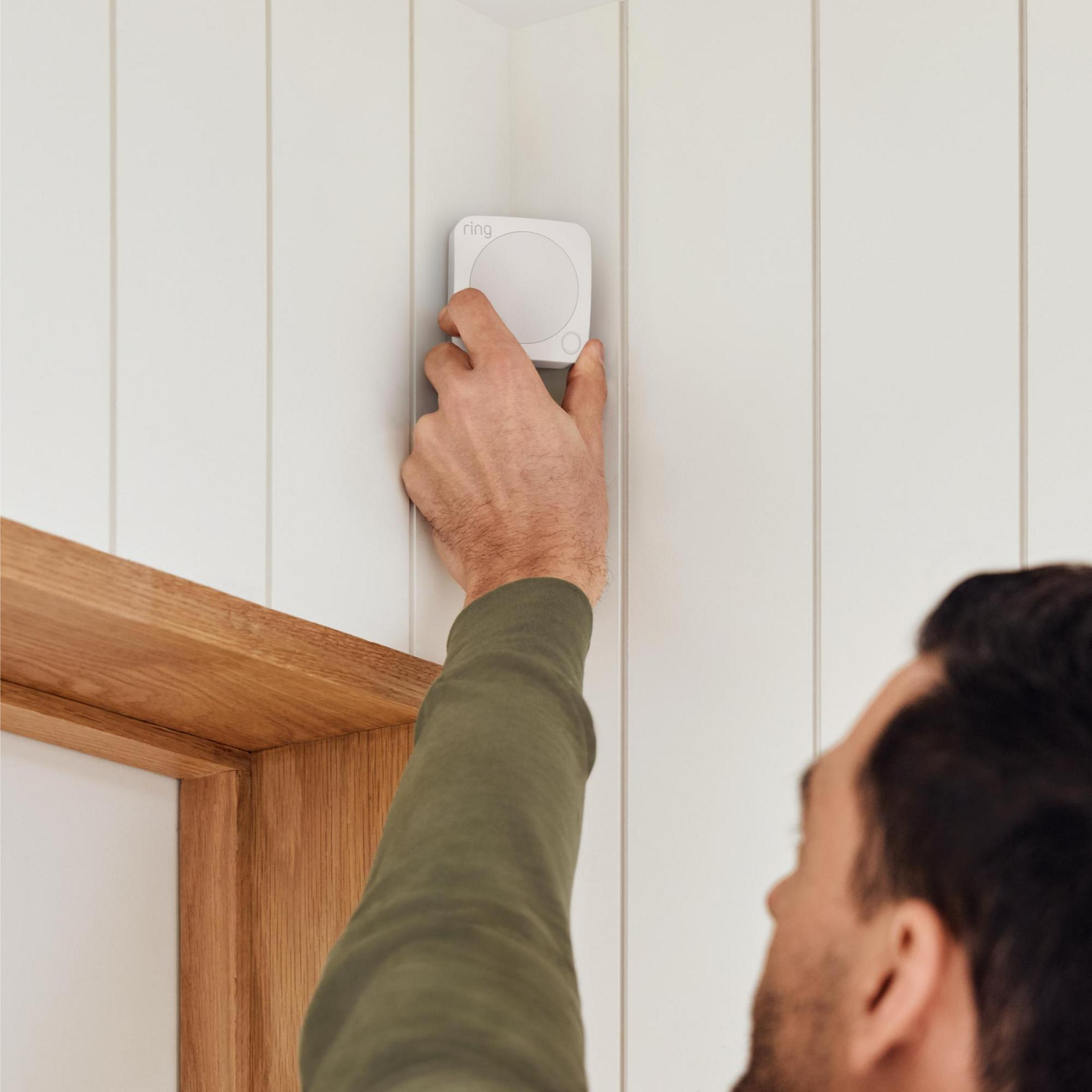 Una persona está instalando un dispositivo blanco con el logo de un anillo en una pared blanca junto a un marco de puerta de madera.