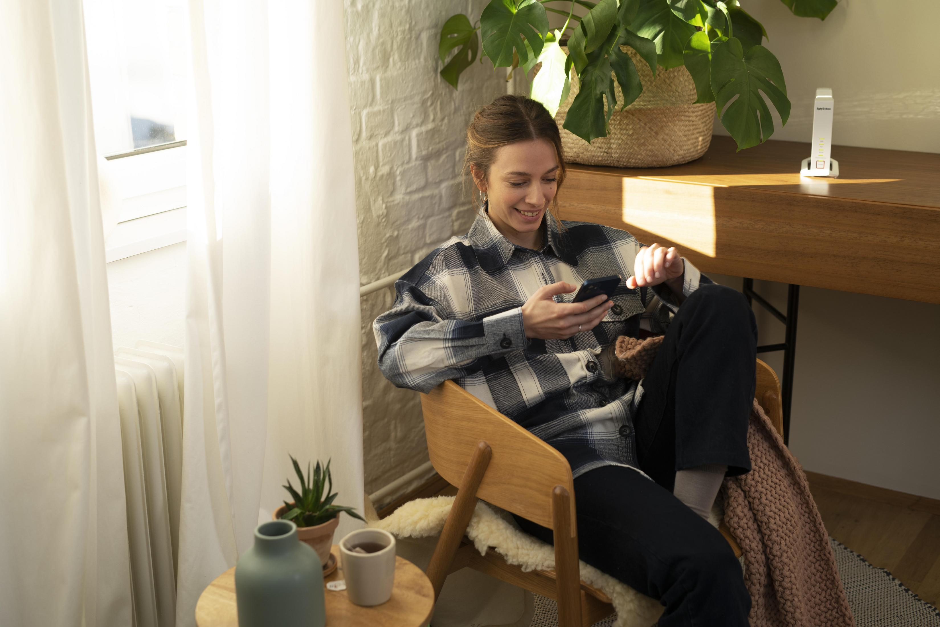 Kobieta w koszuli w kratę, siedząca, używająca smartfona, w pobliżu okna i roślin.