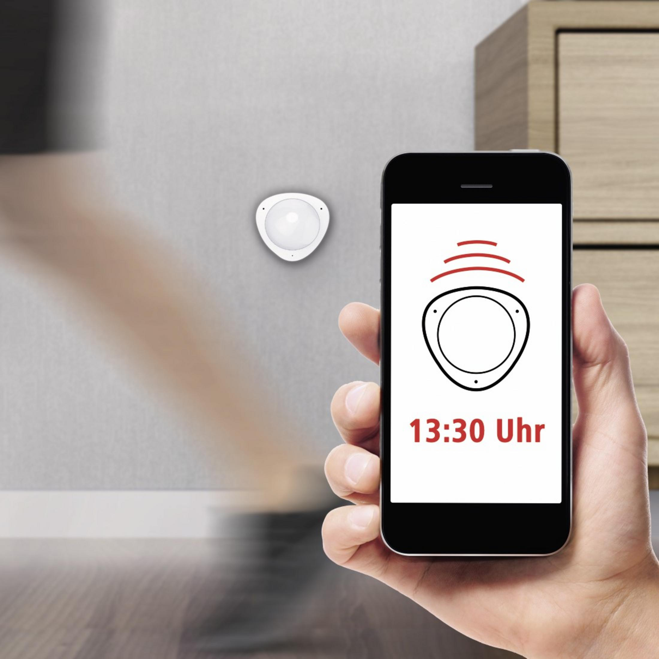 Un sensor blanco en una pared y un teléfono que muestra una interfaz de sensor.